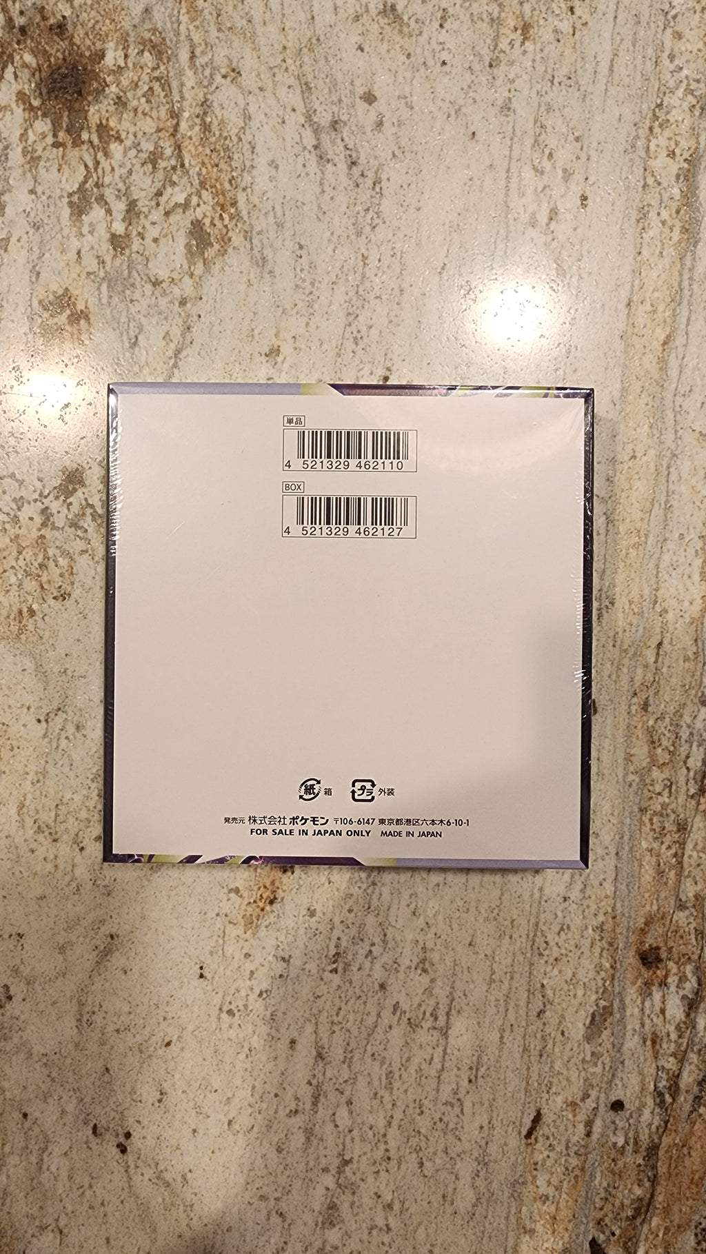 The back of a square white product box or package, showing barcode labels and small printed text, placed on a mottled beige and brown stone surface.