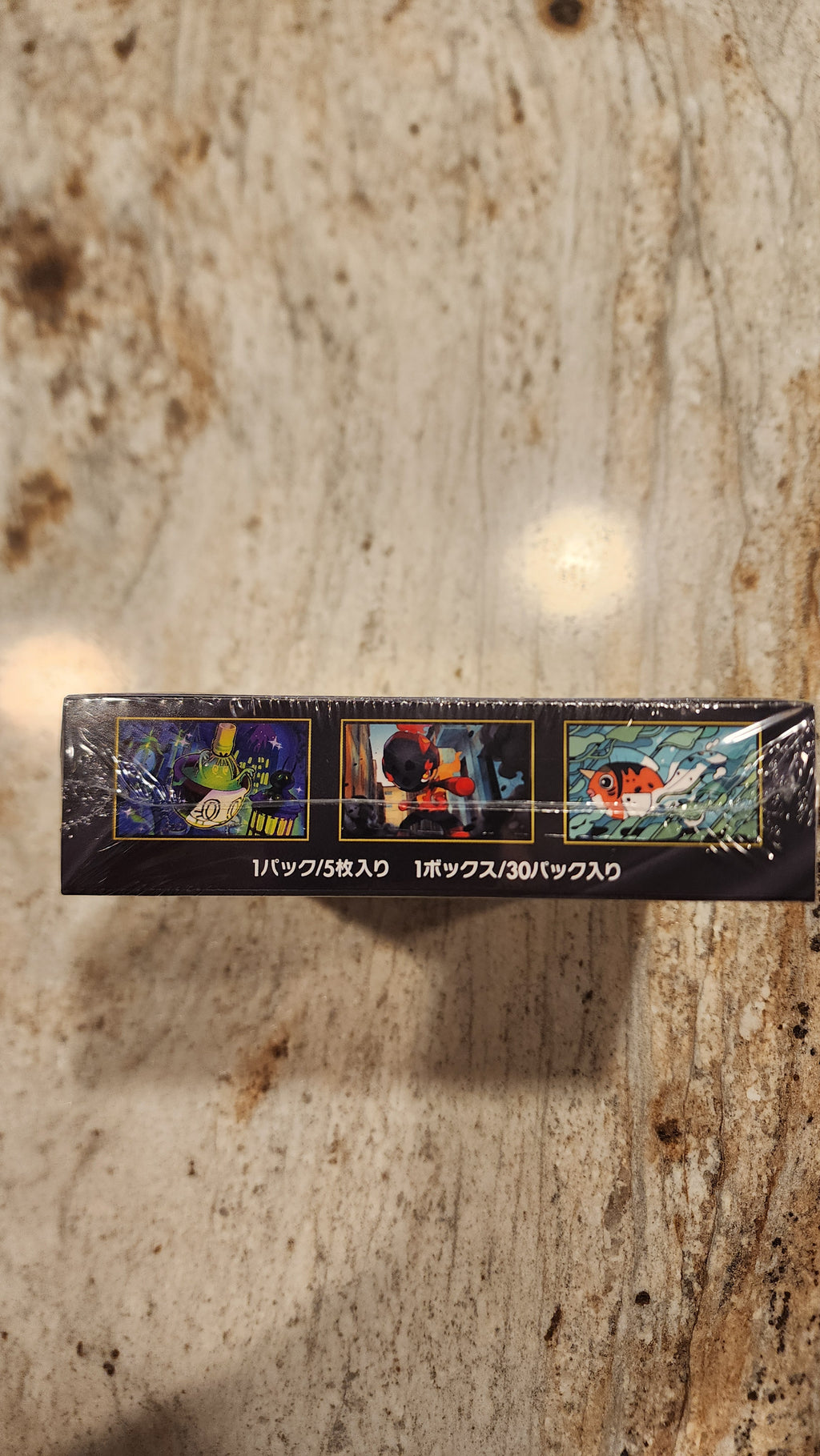 A tall, narrow black product box wrapped in clear plastic, standing vertically on a beige and brown marbled countertop. The box features three small illustrated panels with colorful scenes and vertical white text on the right side.