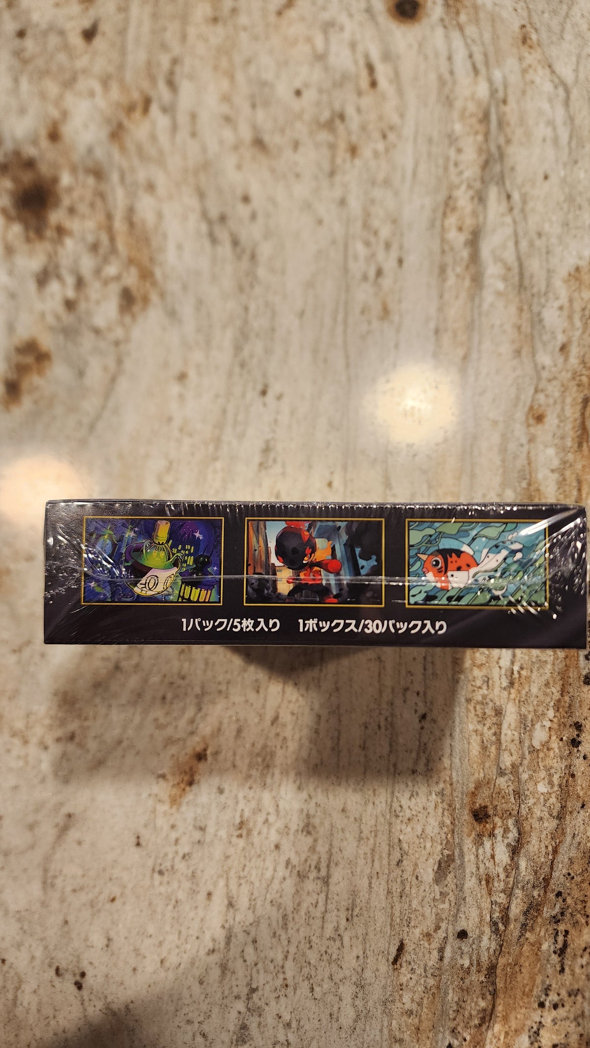 A tall, narrow black product box wrapped in clear plastic, standing vertically on a beige and brown marbled countertop. The box features three small illustrated panels with colorful scenes and vertical white text on the right side.