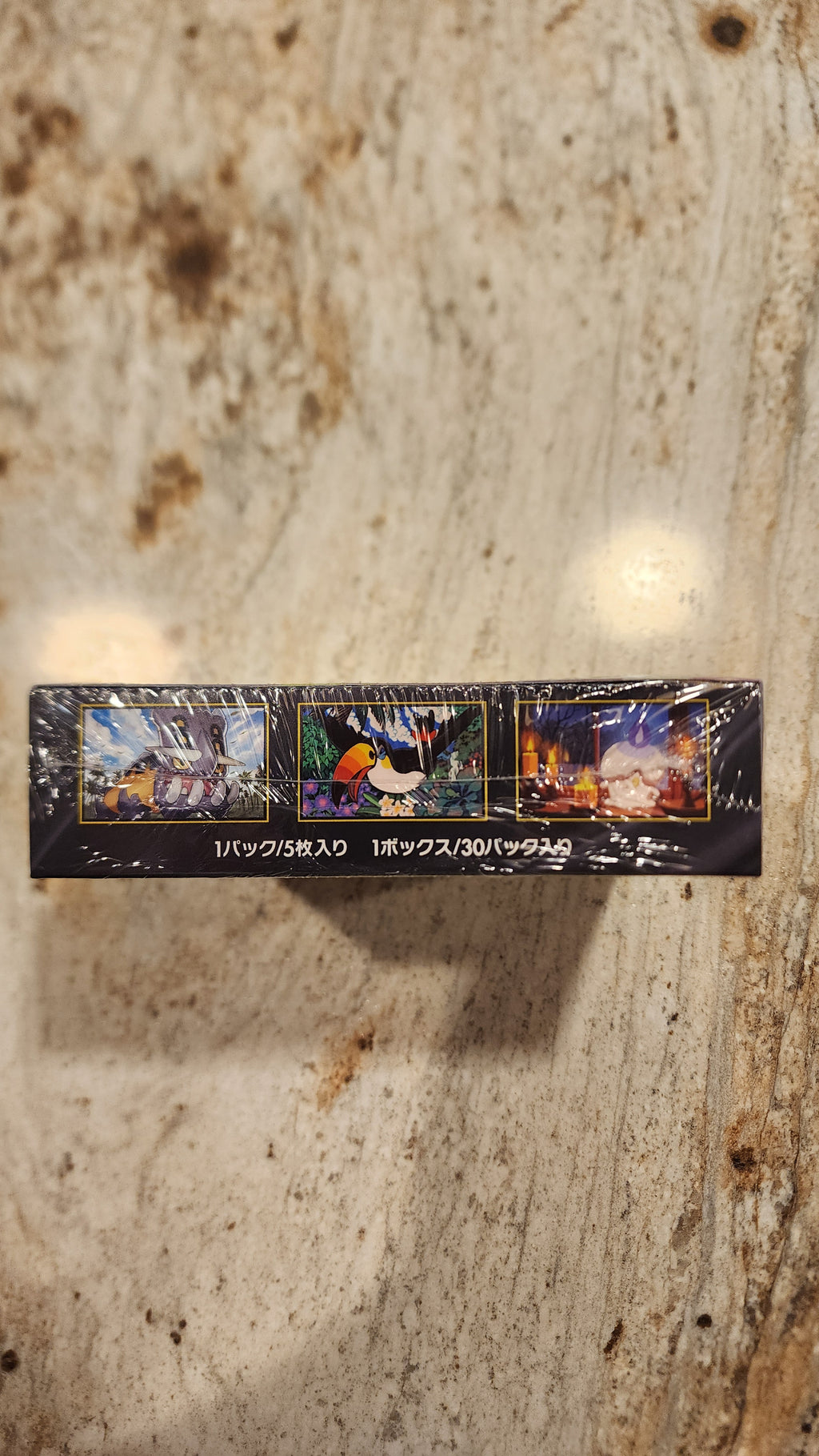 A slim rectangular black product box wrapped in clear plastic, standing upright on a light beige stone-like surface. The box features three colorful illustrated panels and vertical white text.