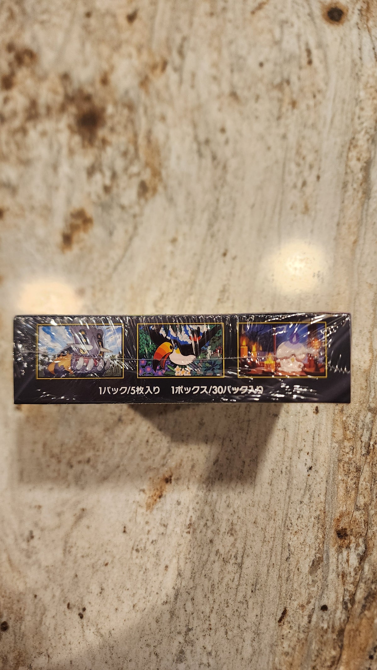 A slim rectangular black product box wrapped in clear plastic, standing upright on a light beige stone-like surface. The box features three colorful illustrated panels and vertical white text.