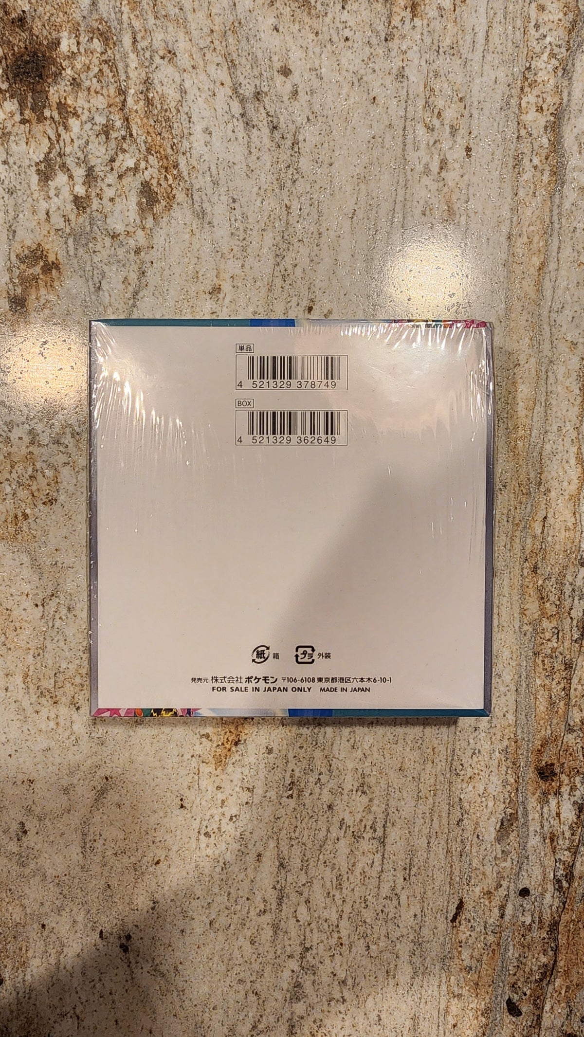 A square white product box wrapped in clear plastic, photographed from above on a mottled beige stone surface. The box has a barcode and small printed text on the front.