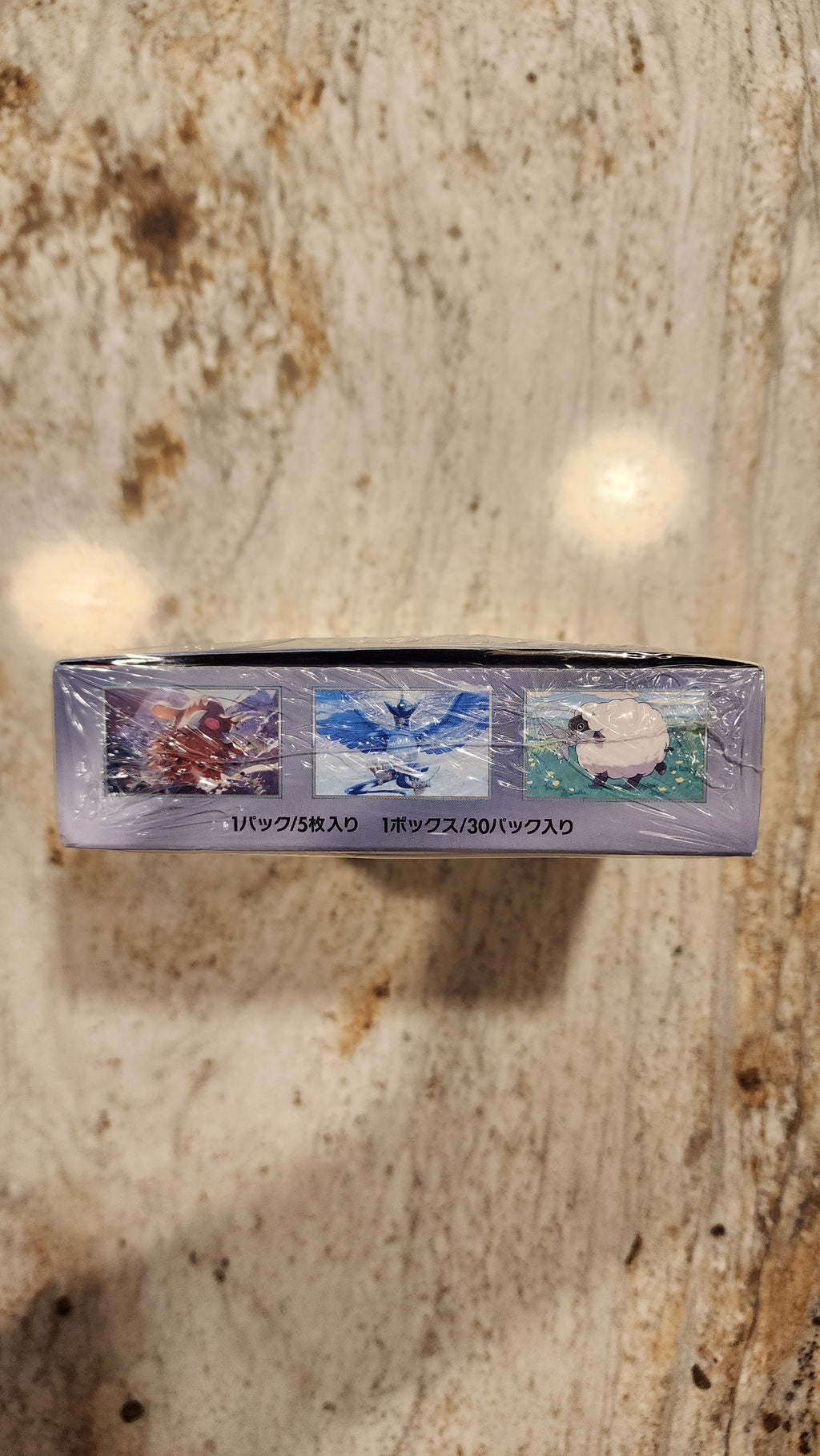 A sealed, shrink-wrapped collectible card box for Pokémon stands vertically on a beige and brown speckled stone surface.
