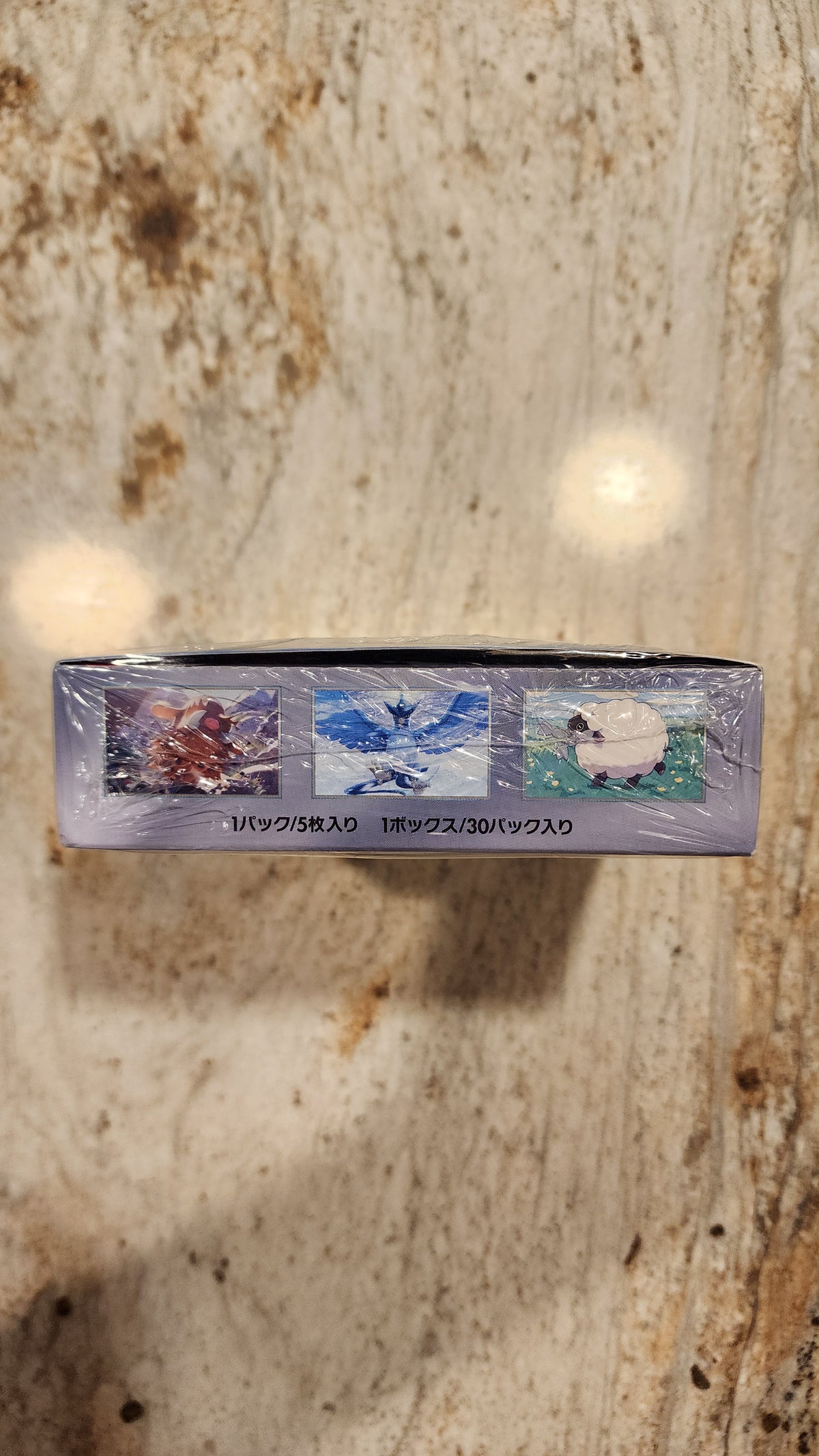 A sealed, shrink-wrapped collectible card box for Pokémon stands vertically on a beige and brown speckled stone surface.