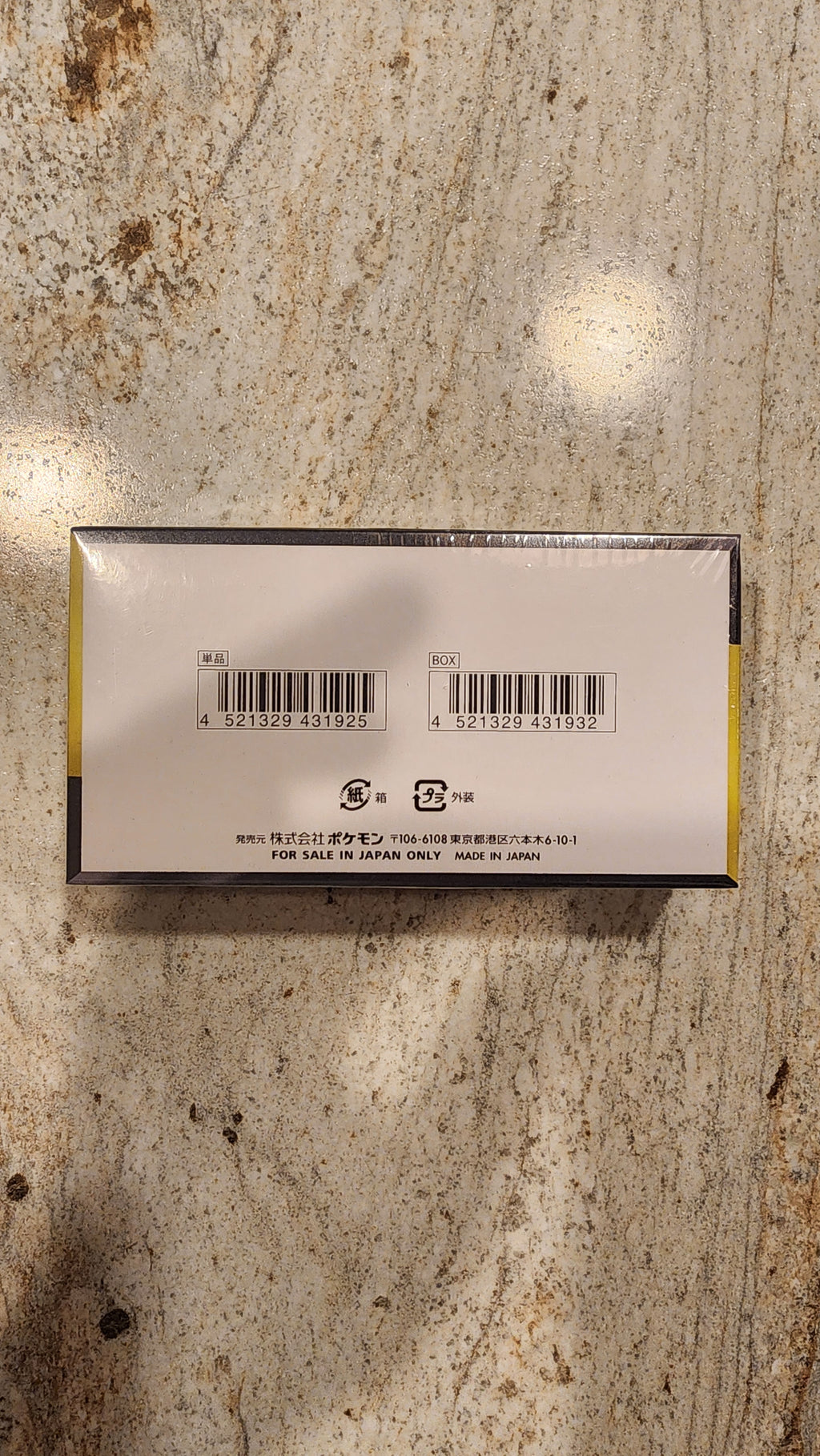 The back of a rectangular product box wrapped in clear plastic, showing barcodes and text, placed on a textured beige and brown stone surface.