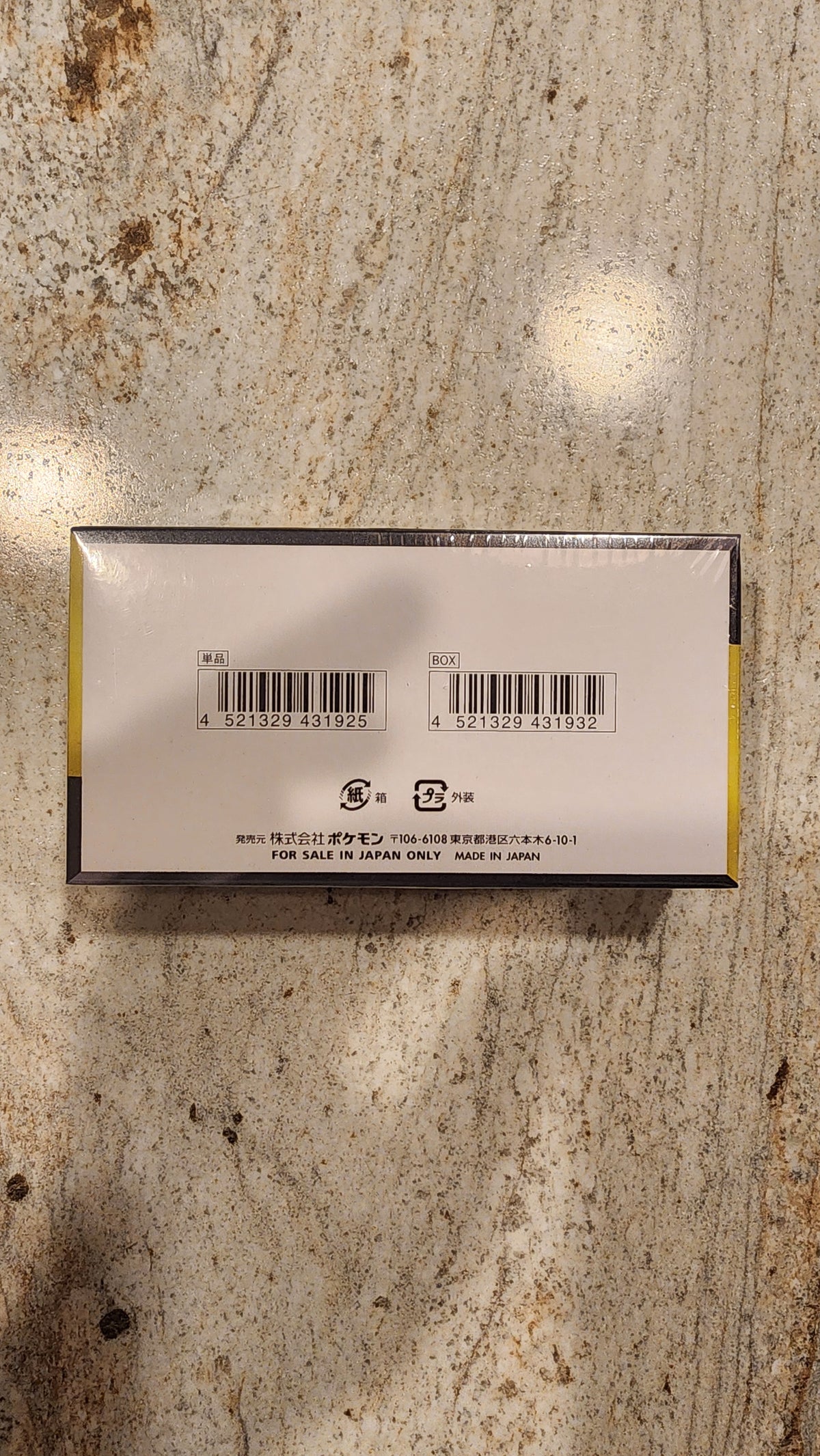 The back of a rectangular product box wrapped in clear plastic, showing barcodes and text, placed on a textured beige and brown stone surface.