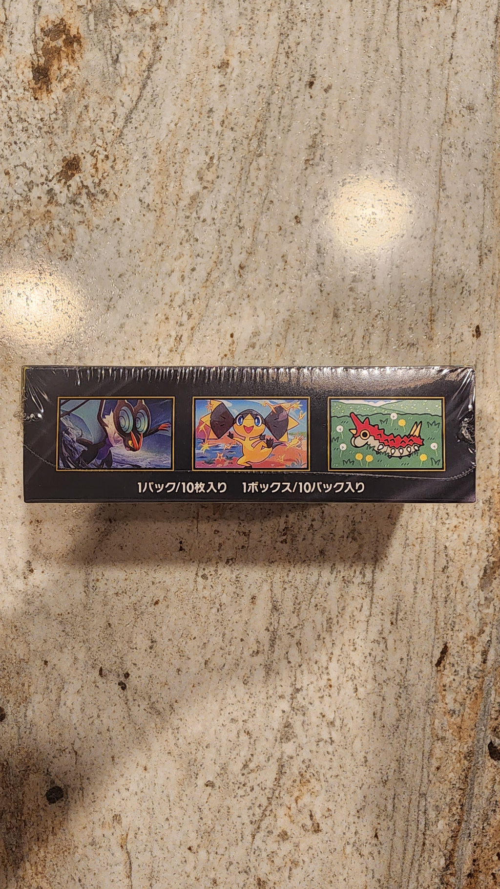 A tall, narrow black product box wrapped in clear plastic, standing upright on a beige stone surface. The box features three colorful illustrated panels and vertical Japanese text.