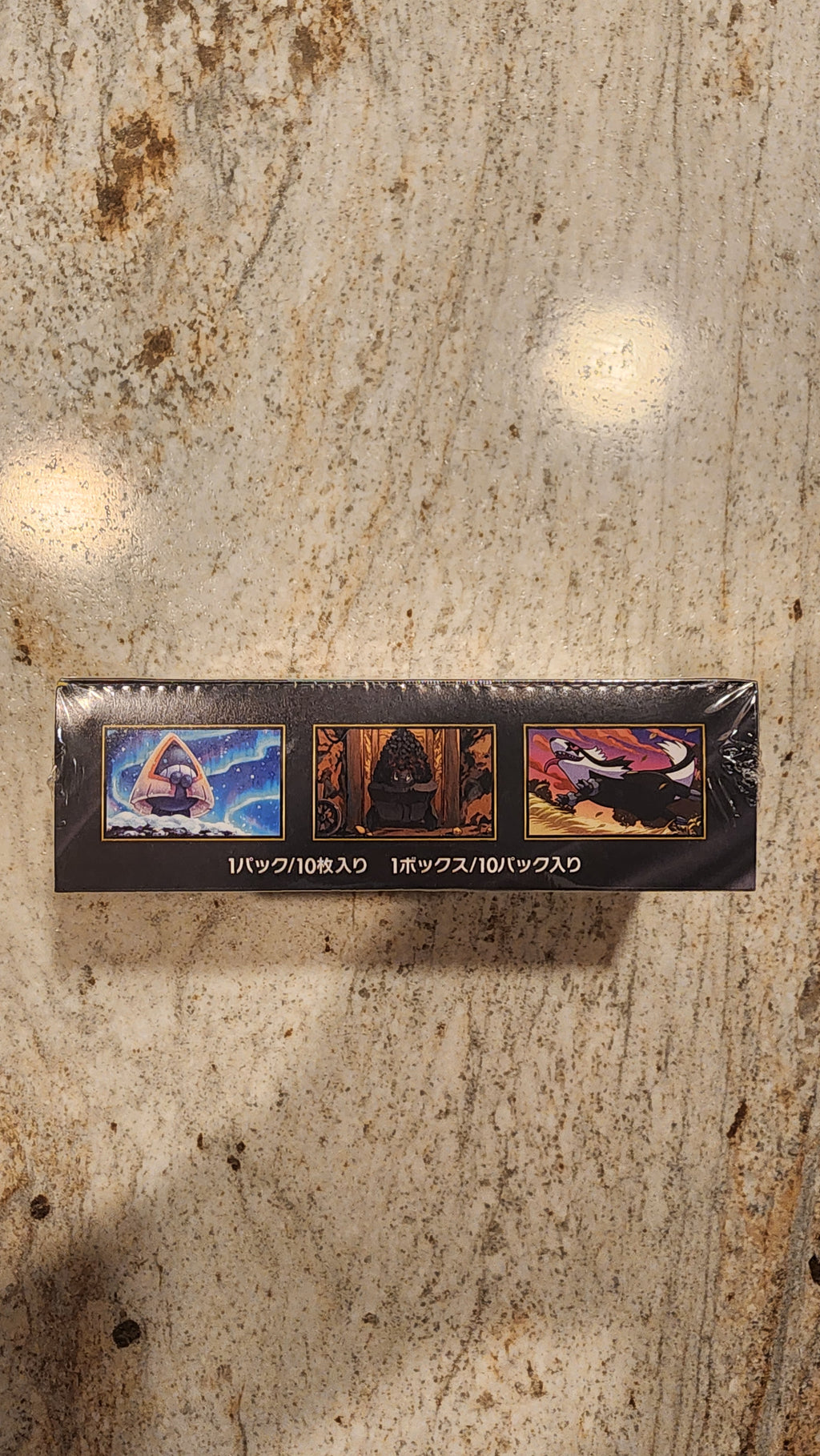 A tall, narrow black product box wrapped in clear plastic, standing upright on a beige stone surface. The box features three colorful illustrated panels and vertical white text reading 'GOLDFINISH MEGA BATTLE'.