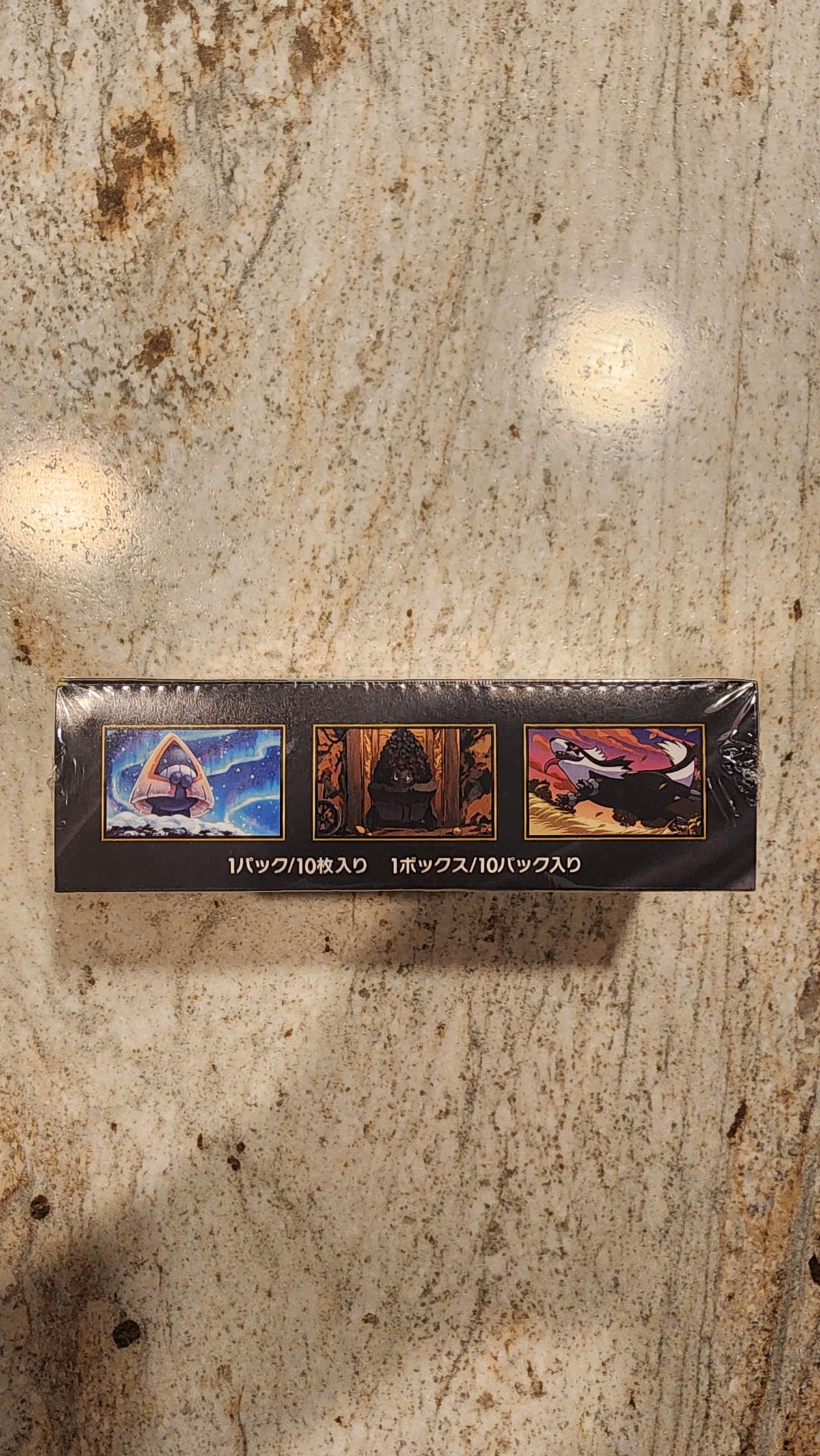 A tall, narrow black product box wrapped in clear plastic, standing upright on a beige stone surface. The box features three colorful illustrated panels and vertical white text reading 'GOLDFINISH MEGA BATTLE'.