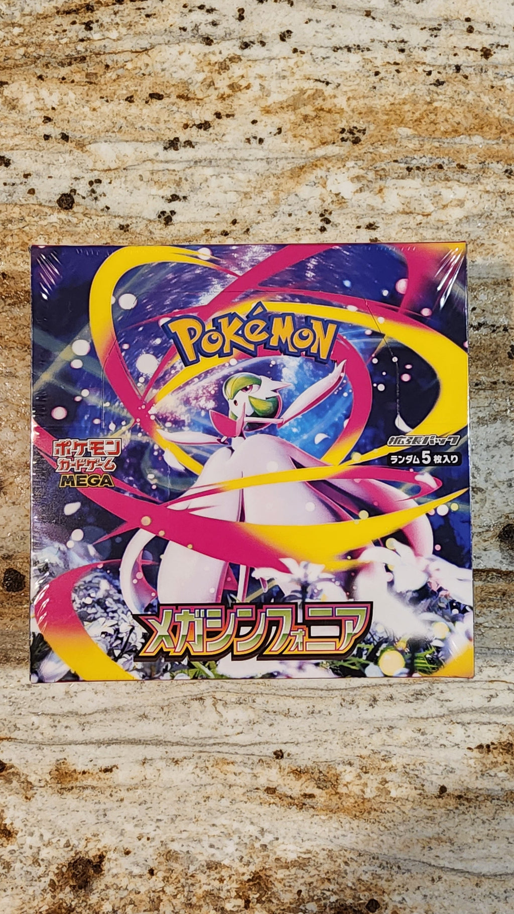 A Pokémon trading card game booster box featuring Mega Symphonia, placed on a textured beige and brown stone surface.