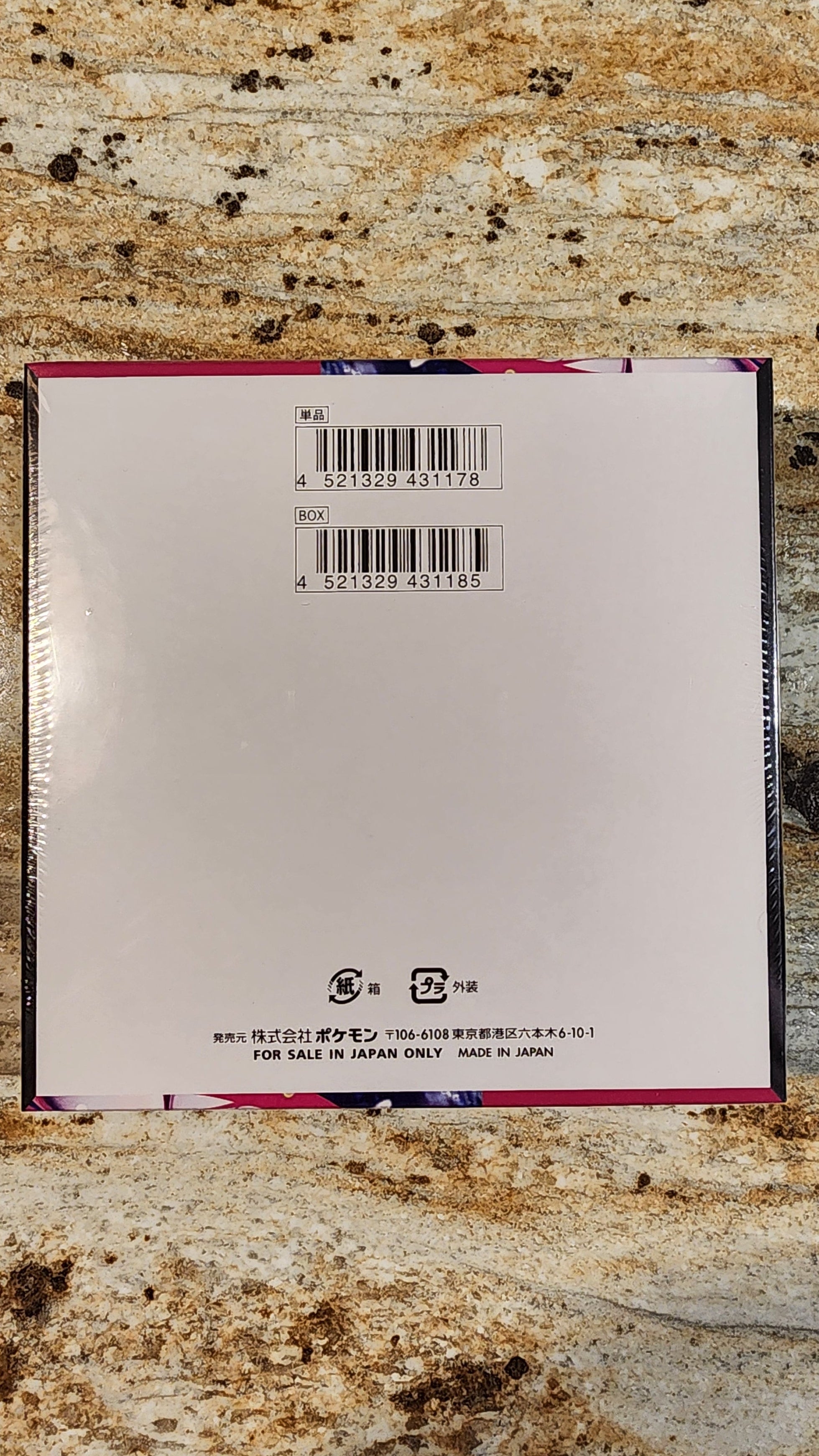 The back of a square white product box wrapped in clear plastic, featuring two barcodes and small printed text near the bottom, placed on a mottled beige and brown stone surface.