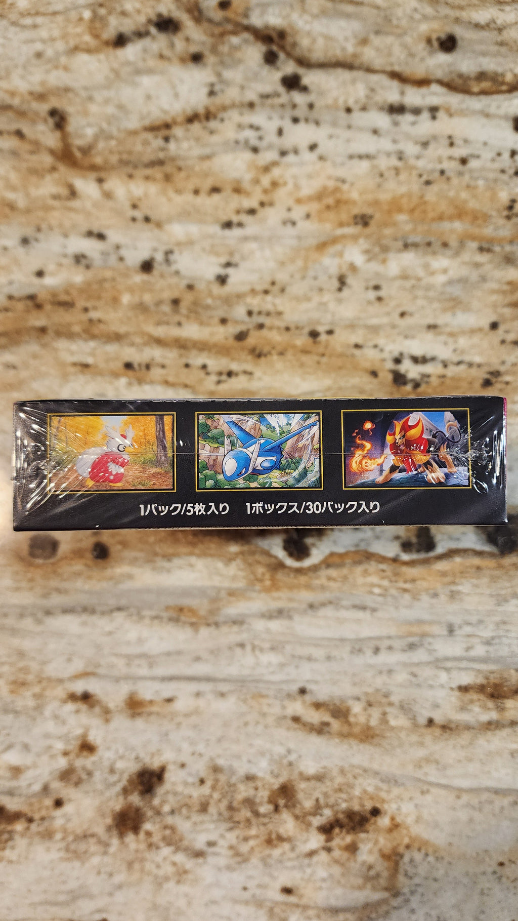 A sealed black box containing three colorful illustrated cards or inserts, photographed on a beige stone surface. The box has Japanese text along the bottom and small fantasy/game-style artwork on each panel.