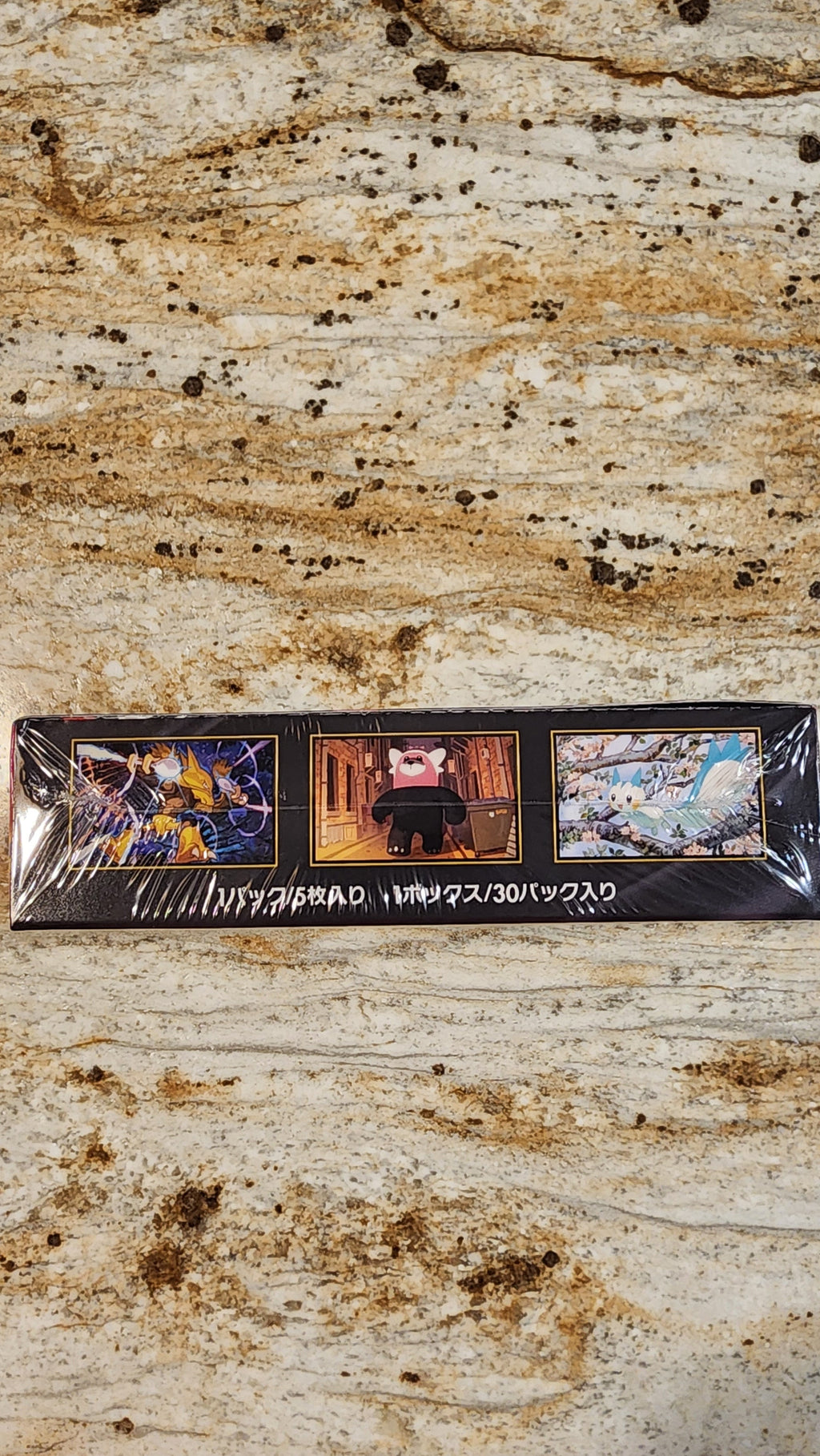 A shrink-wrapped Mega Symphonia box lying on a speckled beige stone surface, with a red fabric edge visible at the bottom.