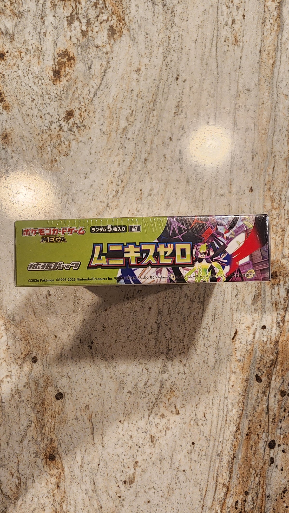 A long, narrow product box with Japanese text and colorful anime-style artwork, standing upright on a mottled beige stone surface.