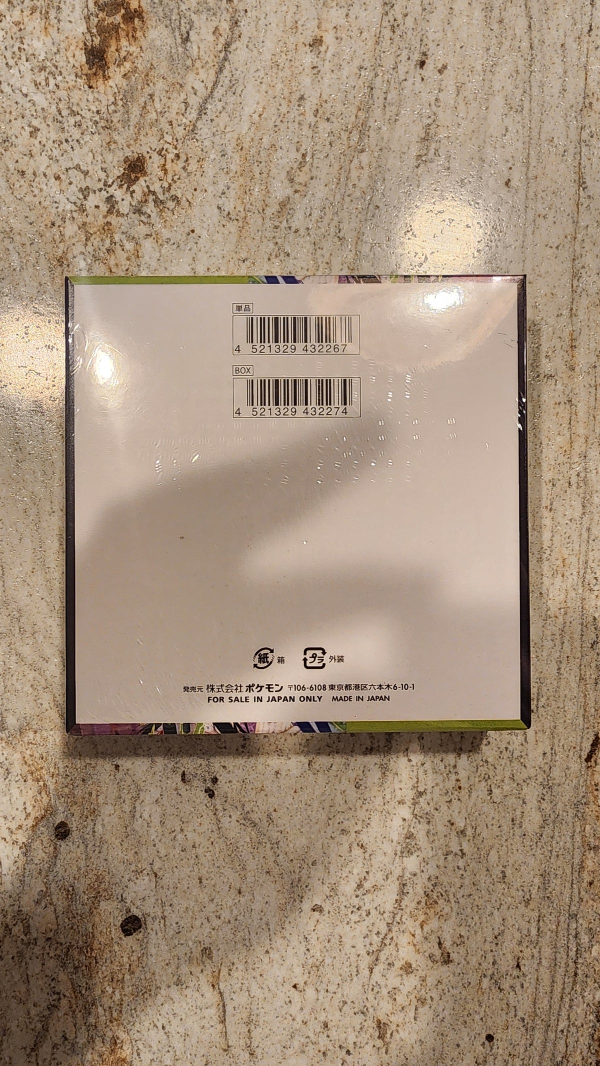 The back of a booster box wrapped in protective plastic, showing a barcode and product information, placed on a textured stone surface.
