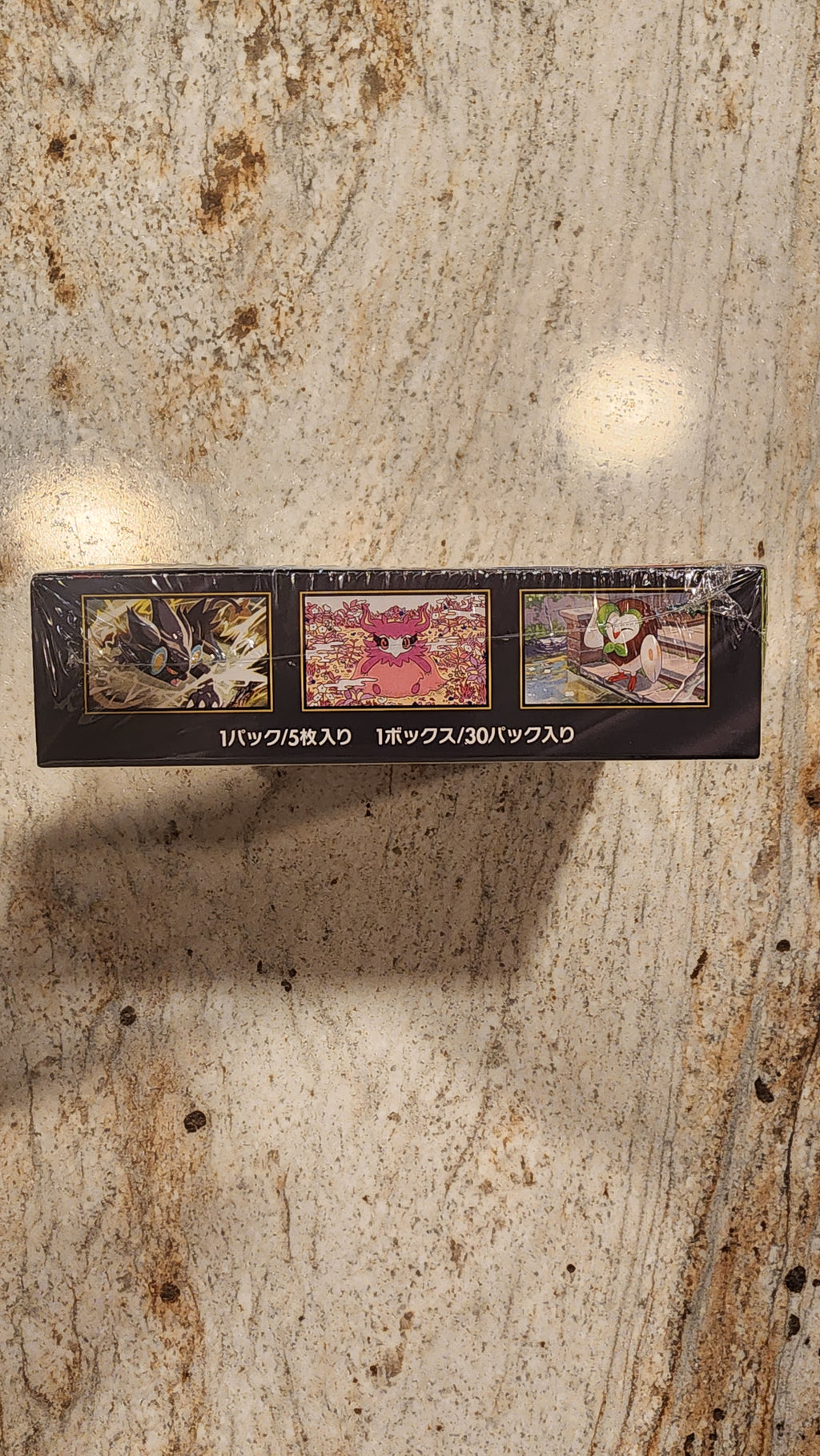 A slim black rectangular product box with three small illustrated panels and vertical Japanese text, standing upright on a beige stone surface with a strong shadow cast to the right.