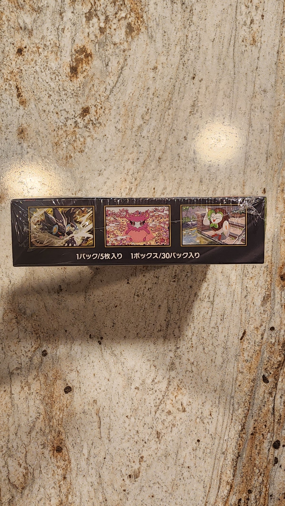 A slim black rectangular product box with three small illustrated panels and vertical Japanese text, standing upright on a beige stone surface with a strong shadow cast to the right.