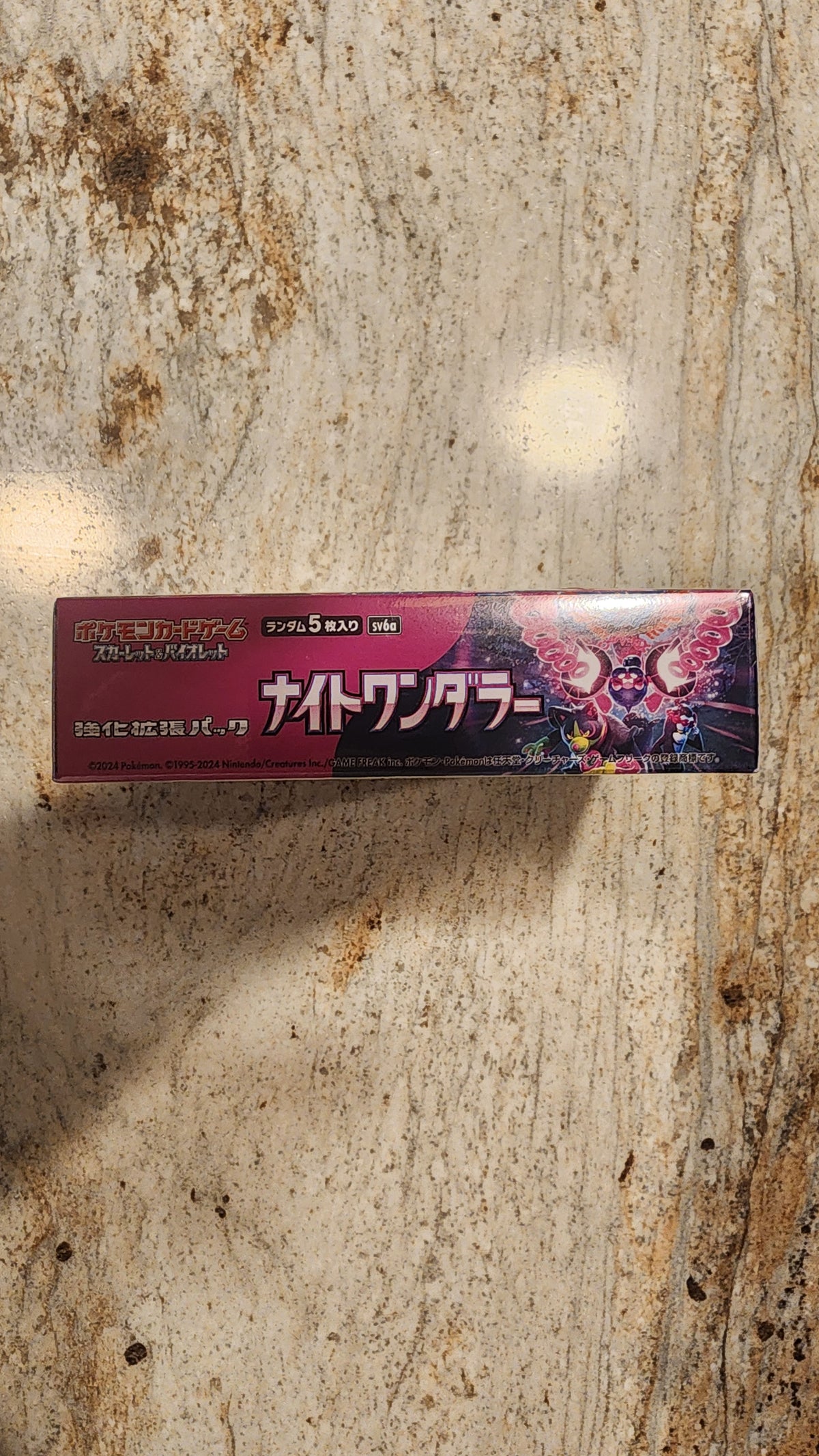 A tall rectangular product box with Japanese text and a dark pink/red design, photographed standing upright on a mottled beige stone-like surface.