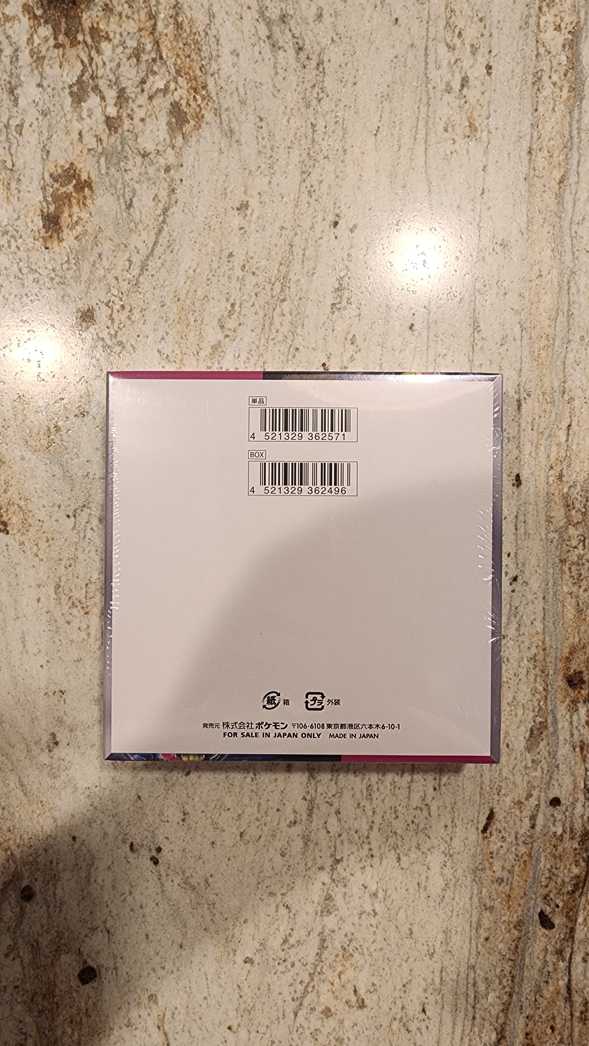 The back of a square CD or DVD case wrapped in clear plastic, showing a barcode and product information, placed on a textured beige stone surface.
