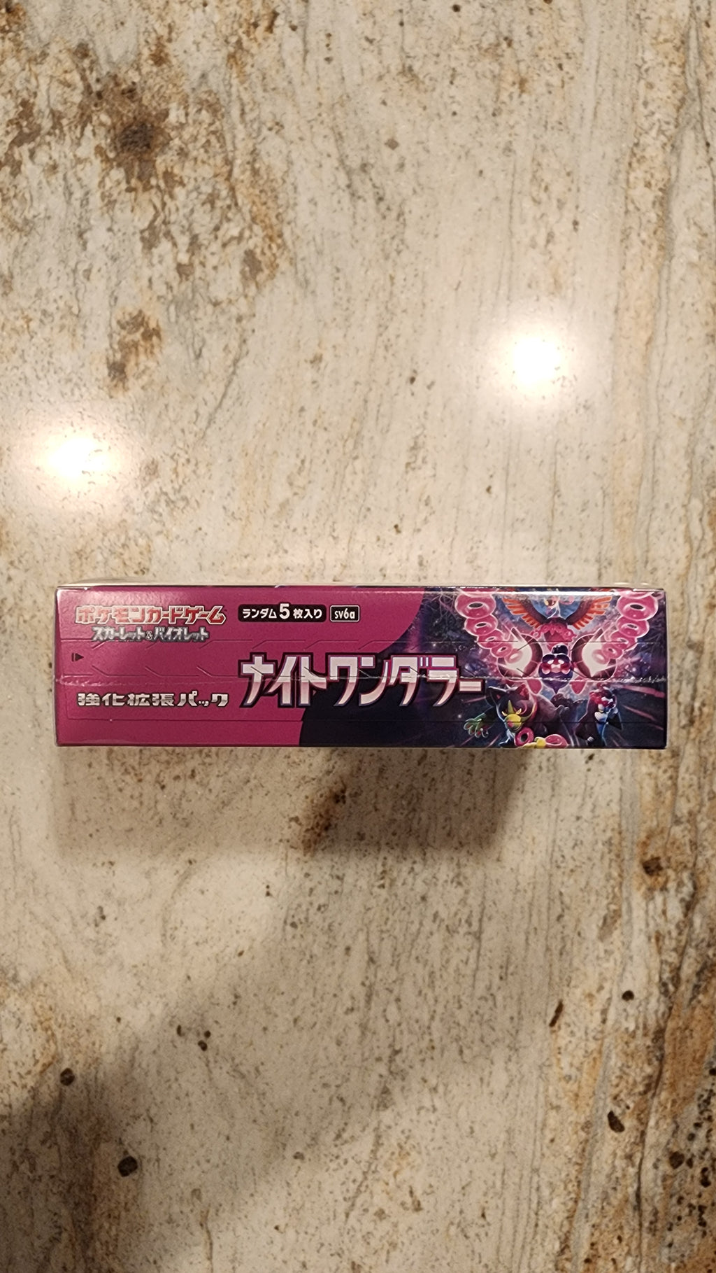 A tall rectangular product box with Japanese text and a dark pink, purple, and black design featuring a dragon illustration, standing on a beige stone surface.