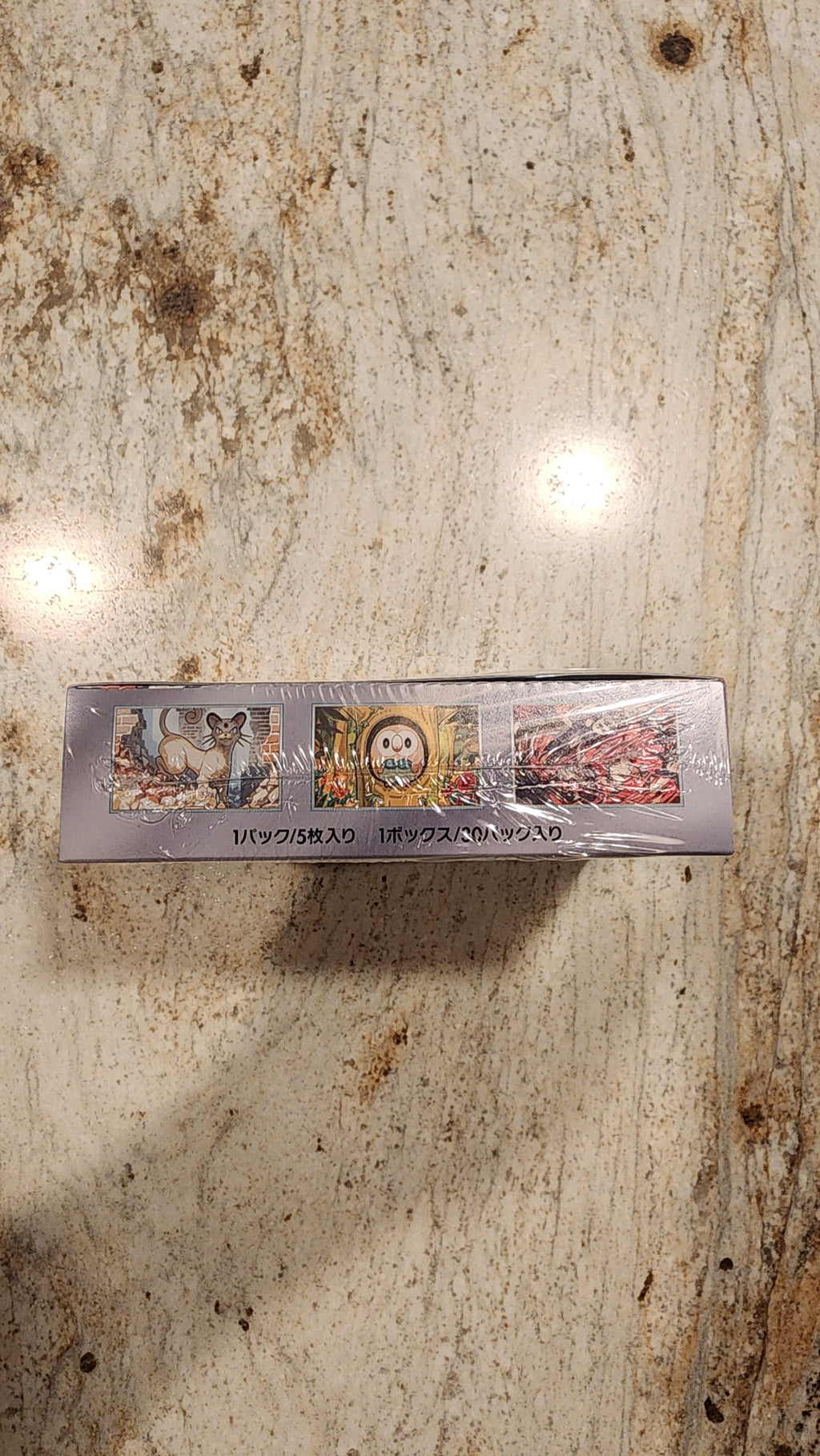 A tall, narrow boxed product wrapped in clear plastic, standing upright on a mottled beige stone-like surface. The box has small printed artwork and vertical text, but the text is not legible enough to identify a brand.