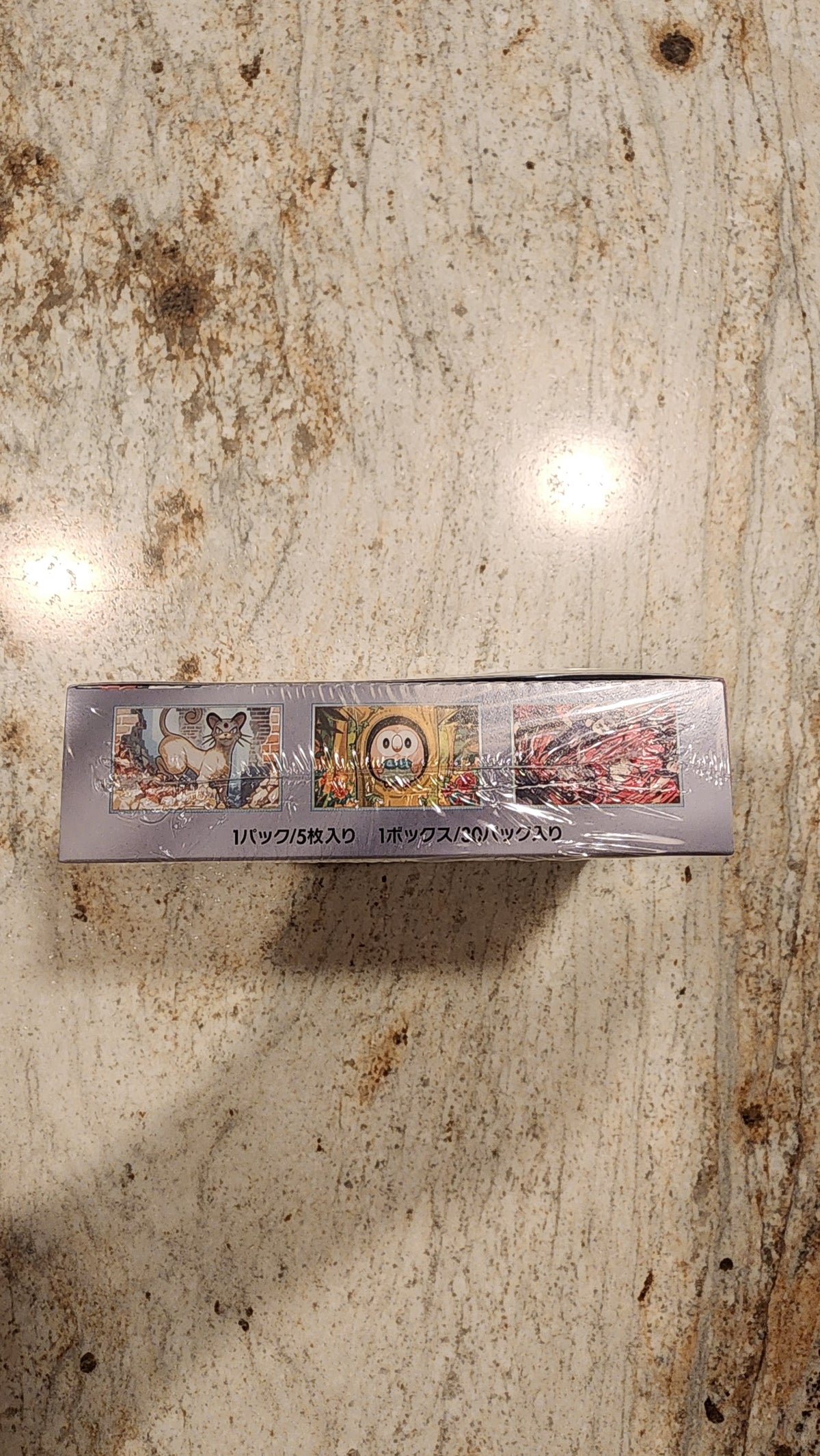 A tall, narrow boxed product wrapped in clear plastic, standing upright on a mottled beige stone-like surface. The box has small printed artwork and vertical text, but the text is not legible enough to identify a brand.