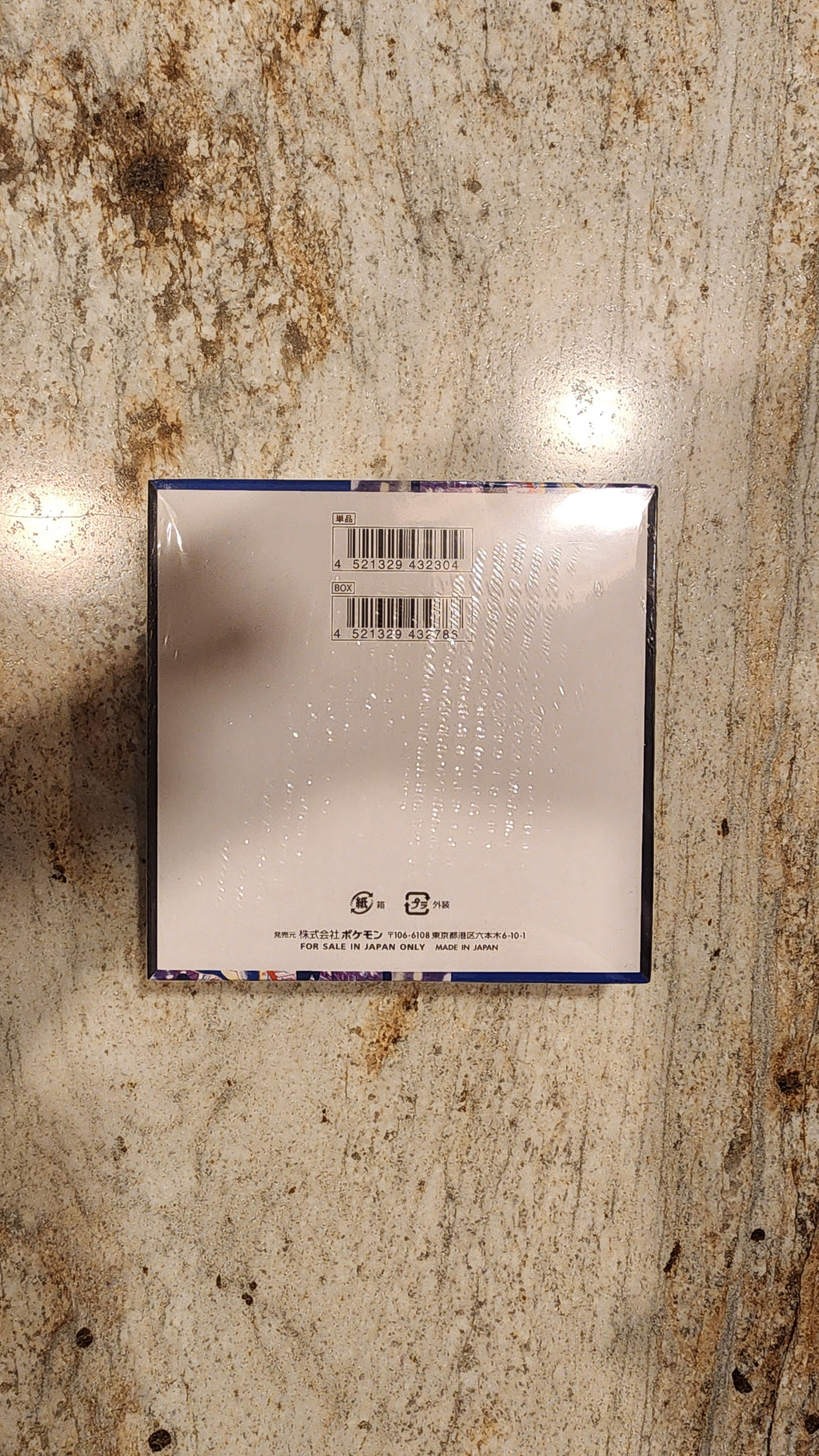 The back of a square product box wrapped in clear plastic, showing a barcode and small printed text, placed on a textured beige stone surface.
