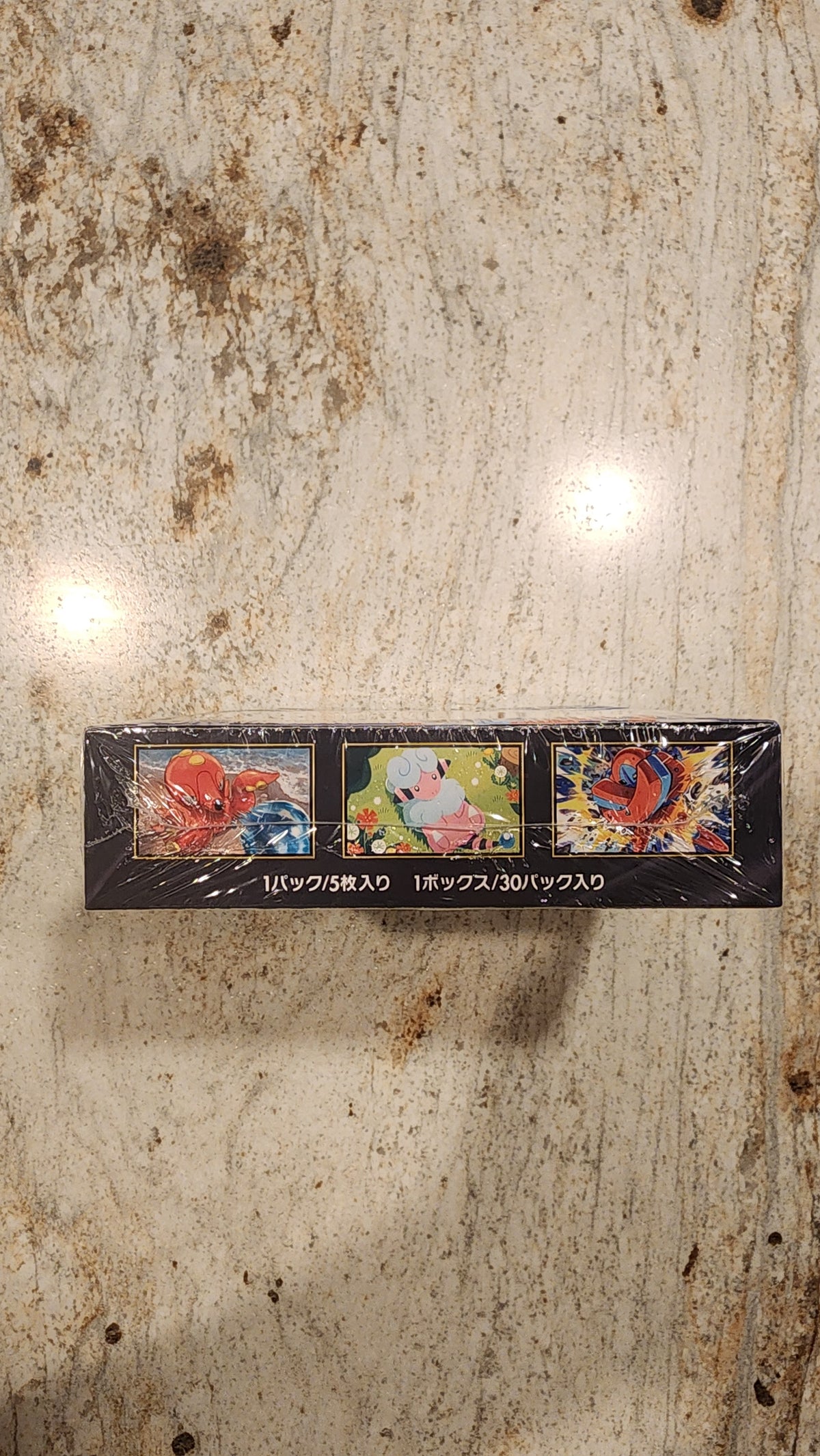 A sealed black rectangular box with three colorful illustrated panels and white vertical text, photographed from above on a mottled beige stone-like surface.