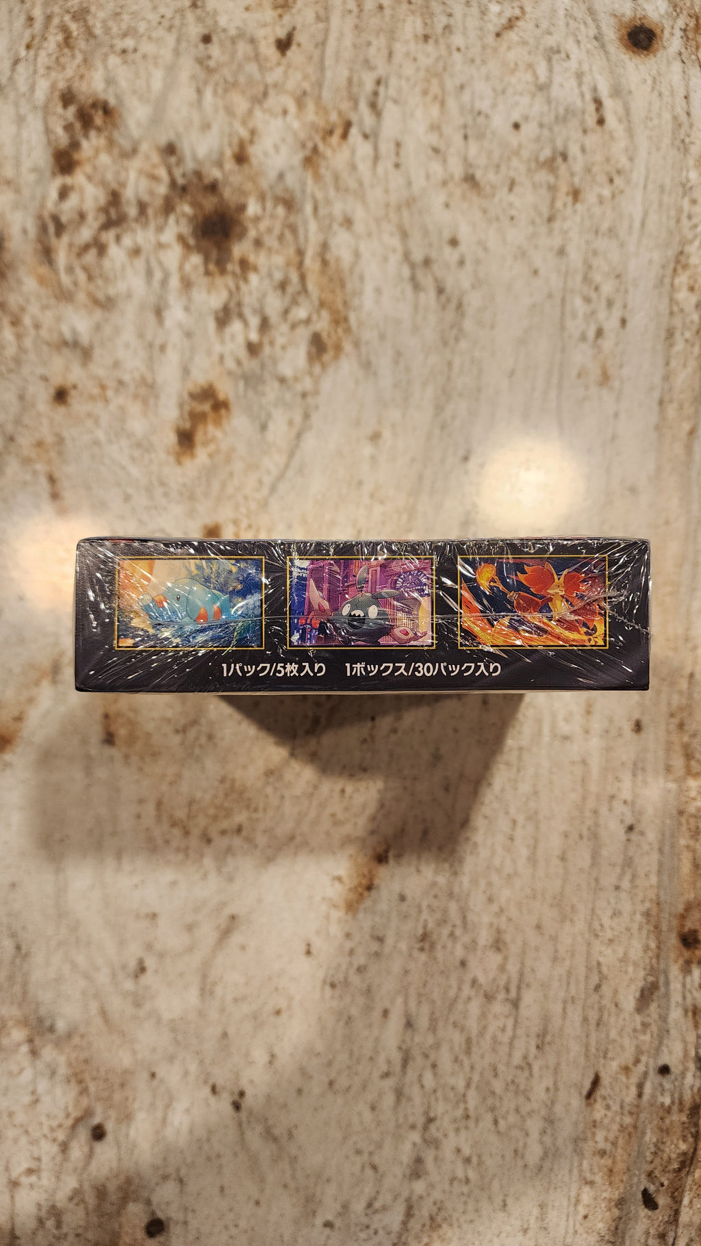 A shrink-wrapped black rectangular box for Pokémon, standing upright on a beige marble-like surface. The box features three colorful card art panels and vertical white text.