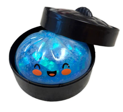 A blue glittery squishy ball toy with a cute cartoon face, presented inside a black round container with its lid propped open, on a white background.