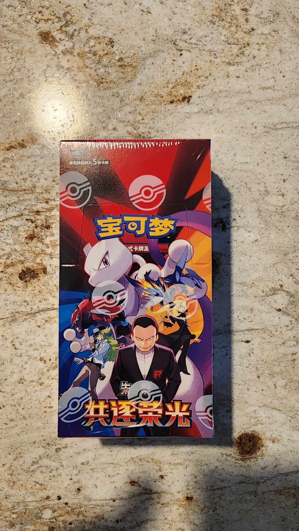 A Pokémon booster pack box featuring colorful character artwork, photographed standing on a mottled stone-like surface.