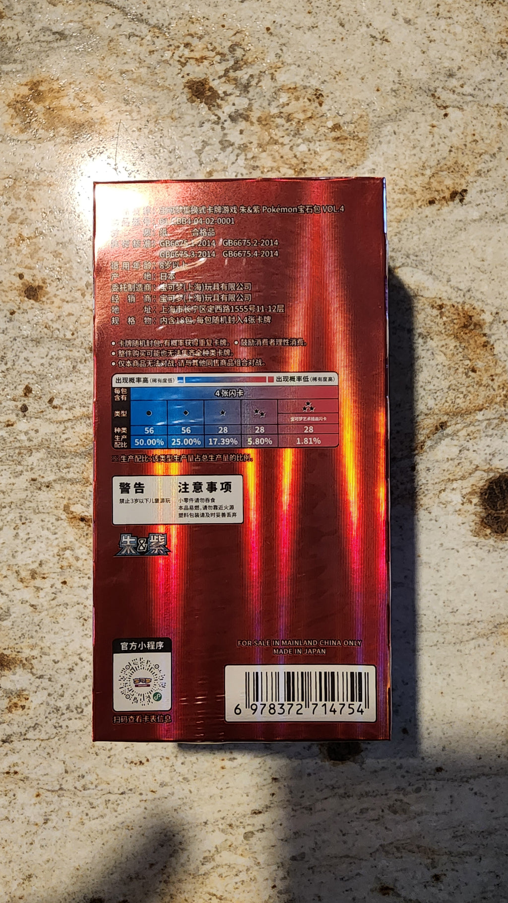 The back of a red metallic box with Chinese text, a blue and red warning label, and a barcode, placed on a speckled beige stone surface.