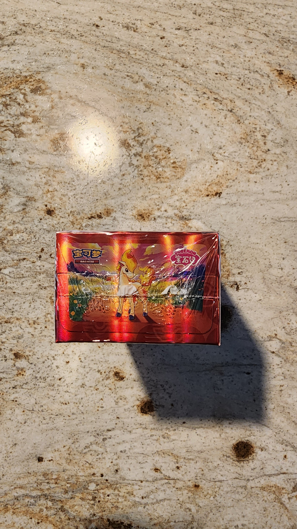 A rectangular red box with colorful illustrated artwork on the front, photographed from above on a textured beige stone surface with a strong shadow cast to the right.