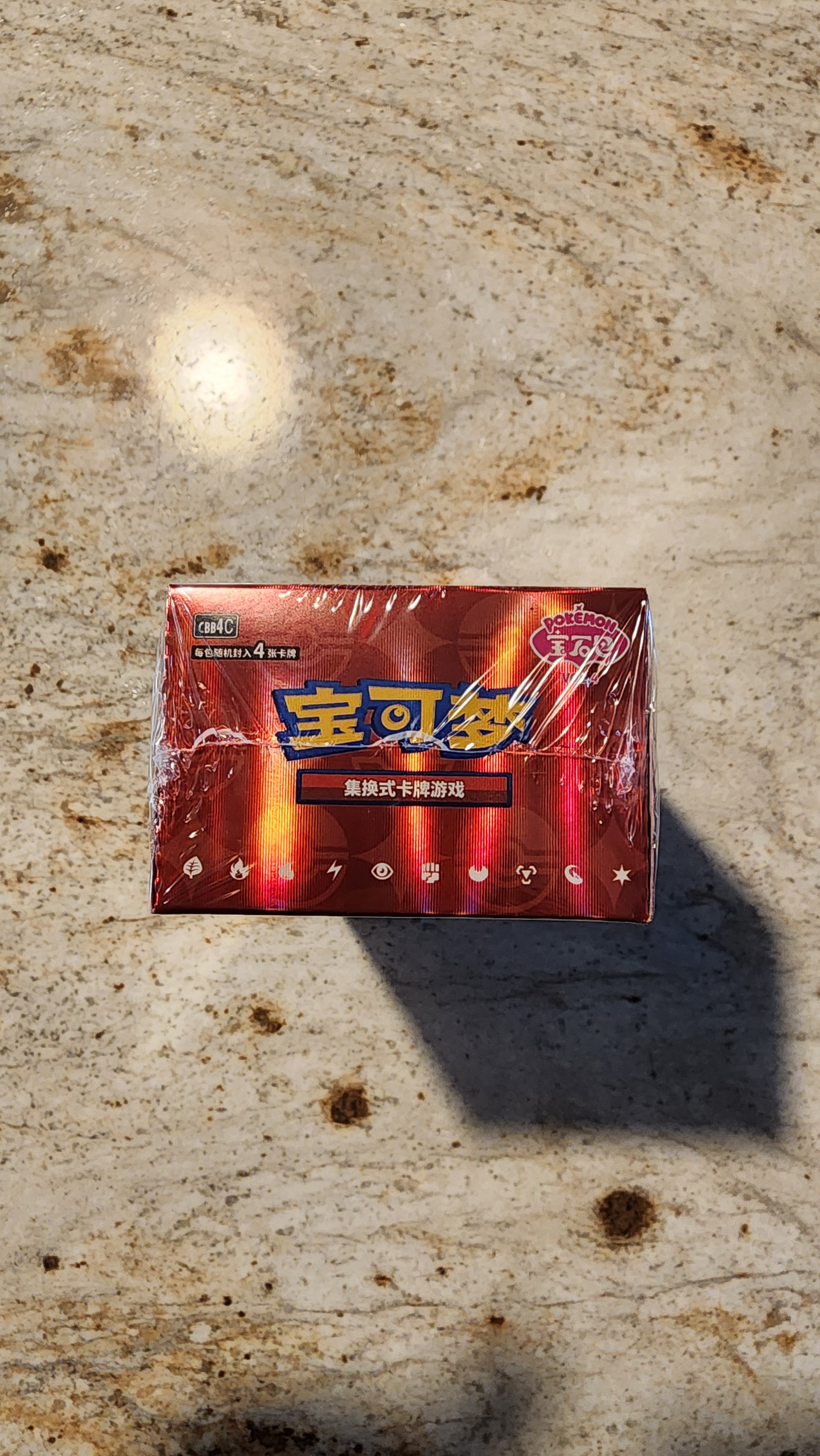 A sealed red and orange box of Chinese trading cards, branded 宝可多 (Bakemono), placed on a beige marble surface with a strong shadow cast to the right.