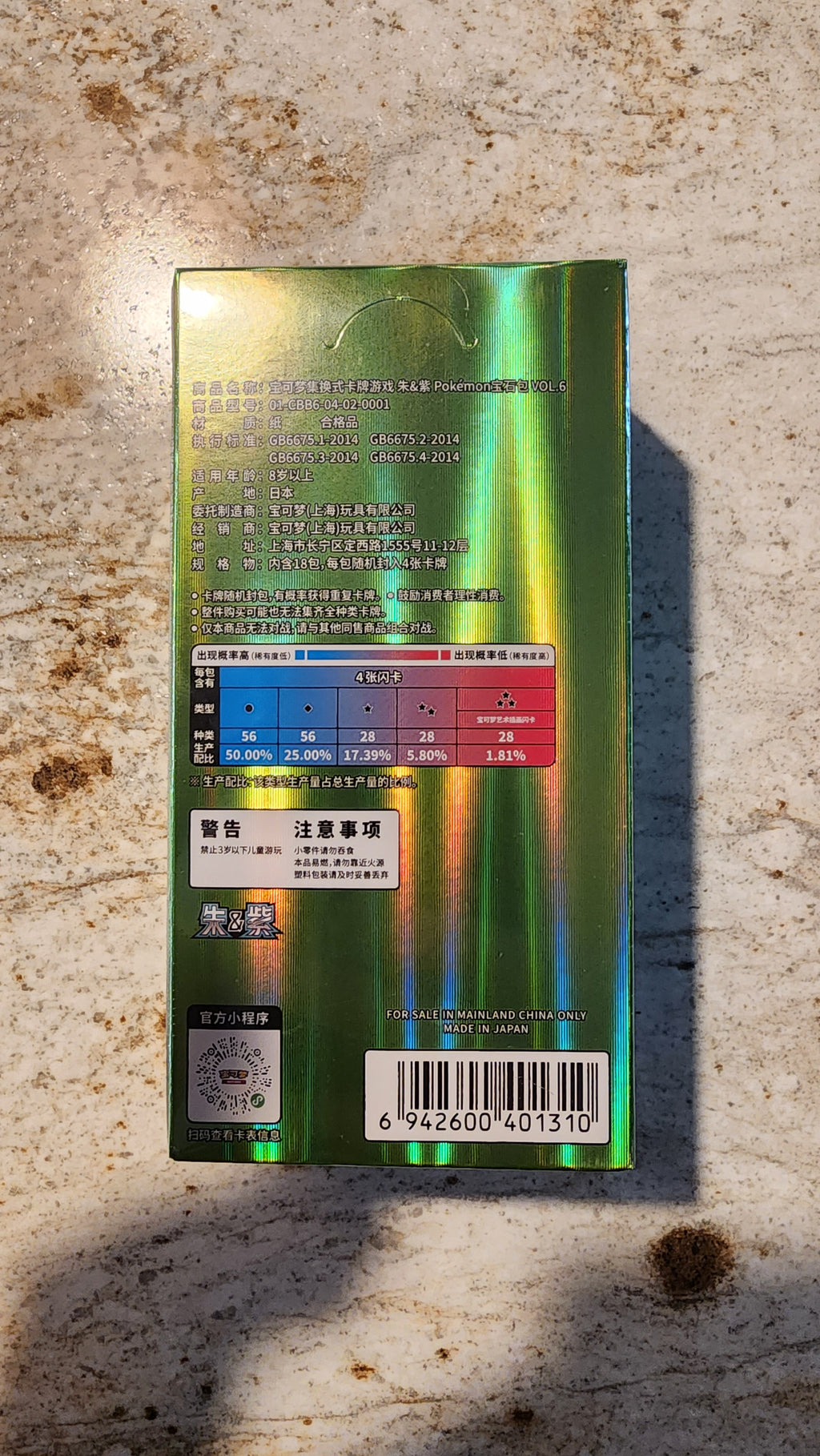 The back of a green holographic box with Chinese text, a color chart, and a barcode, lying on a textured beige stone surface.