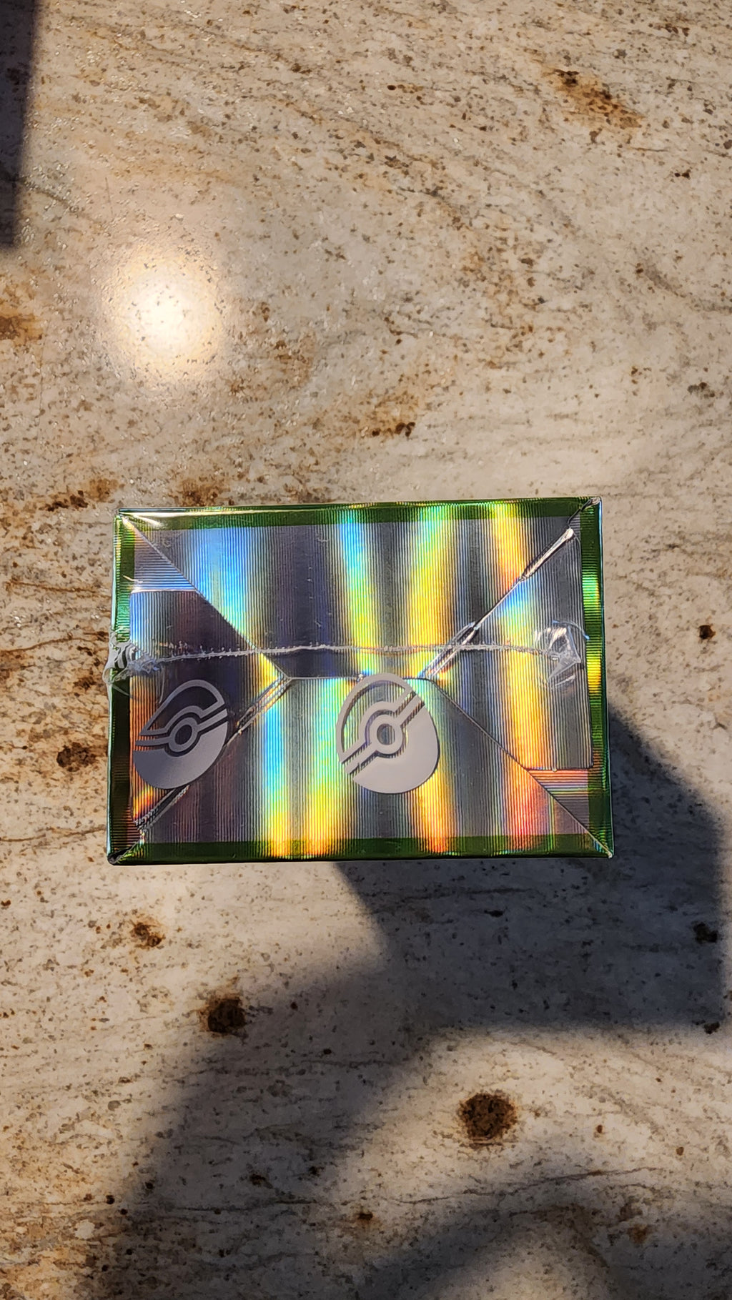 A rectangular holographic card case with a metallic iridescent finish and Pokémon logo, placed on a textured beige stone surface.