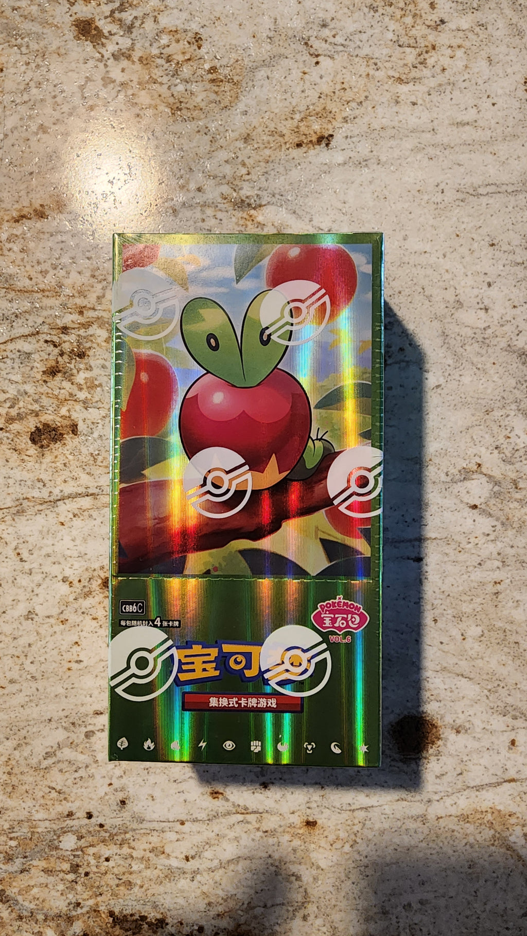 A sealed Pokémon trading card booster pack featuring a shiny holographic green and red Apple Sulfurash design, placed on a textured beige stone surface.