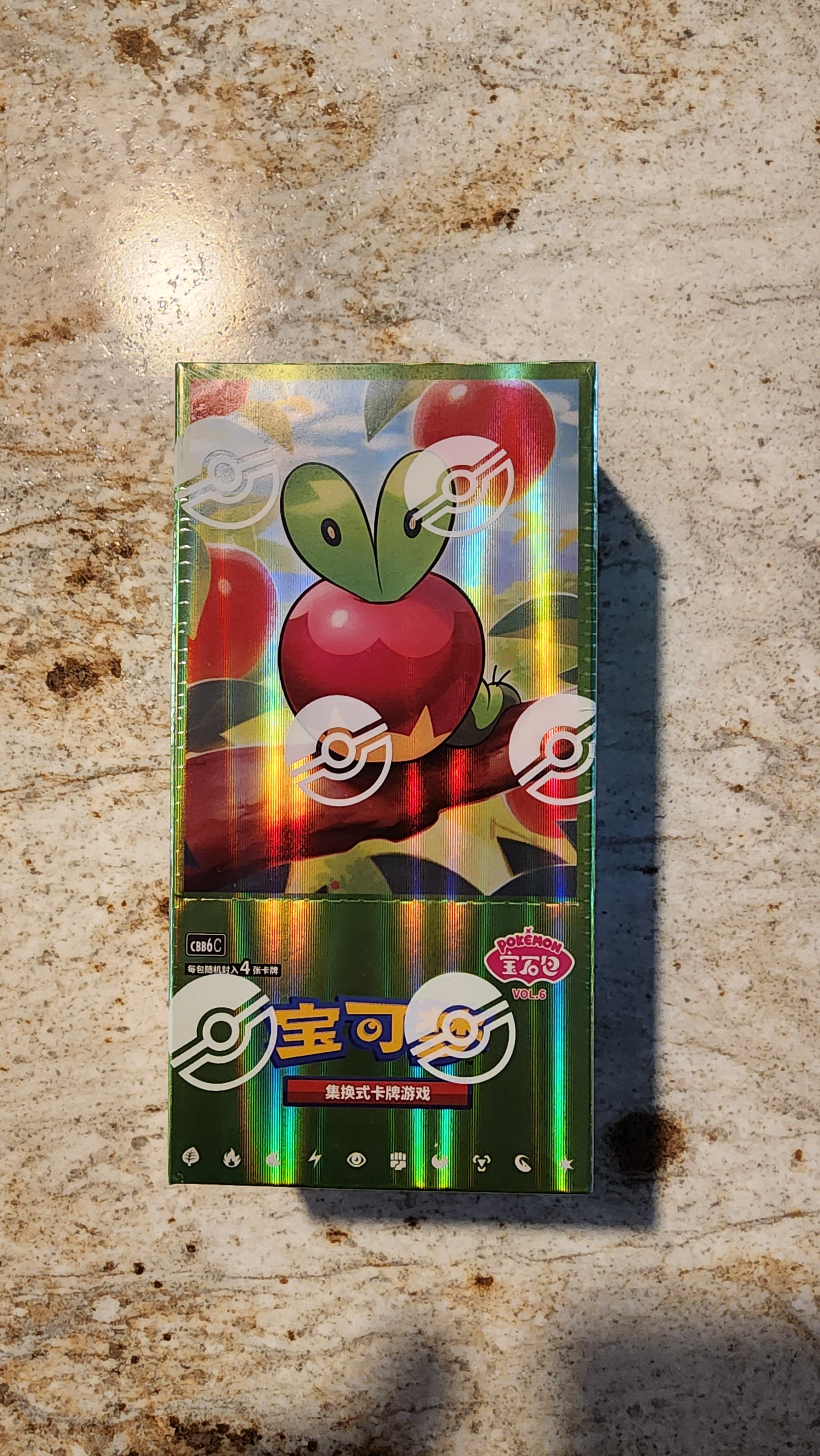 A sealed Pokémon trading card booster pack featuring a shiny holographic green and red Apple Sulfurash design, placed on a textured beige stone surface.