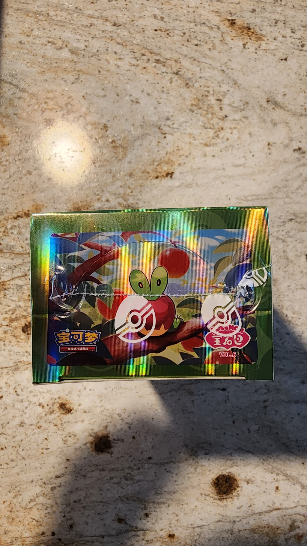 A sealed Pokémon trading card booster pack featuring a green leafy character on a holographic silver background, placed on a light stone surface.