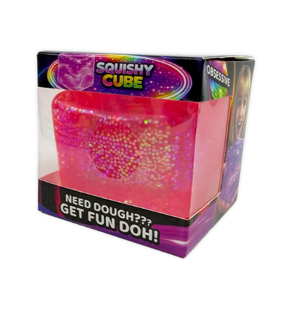 A pink and purple 'Squishy Cube' sensory toy in its original retail packaging, featuring a translucent pink cube filled with gold glitter and a heart-shaped pattern inside a clear plastic box.