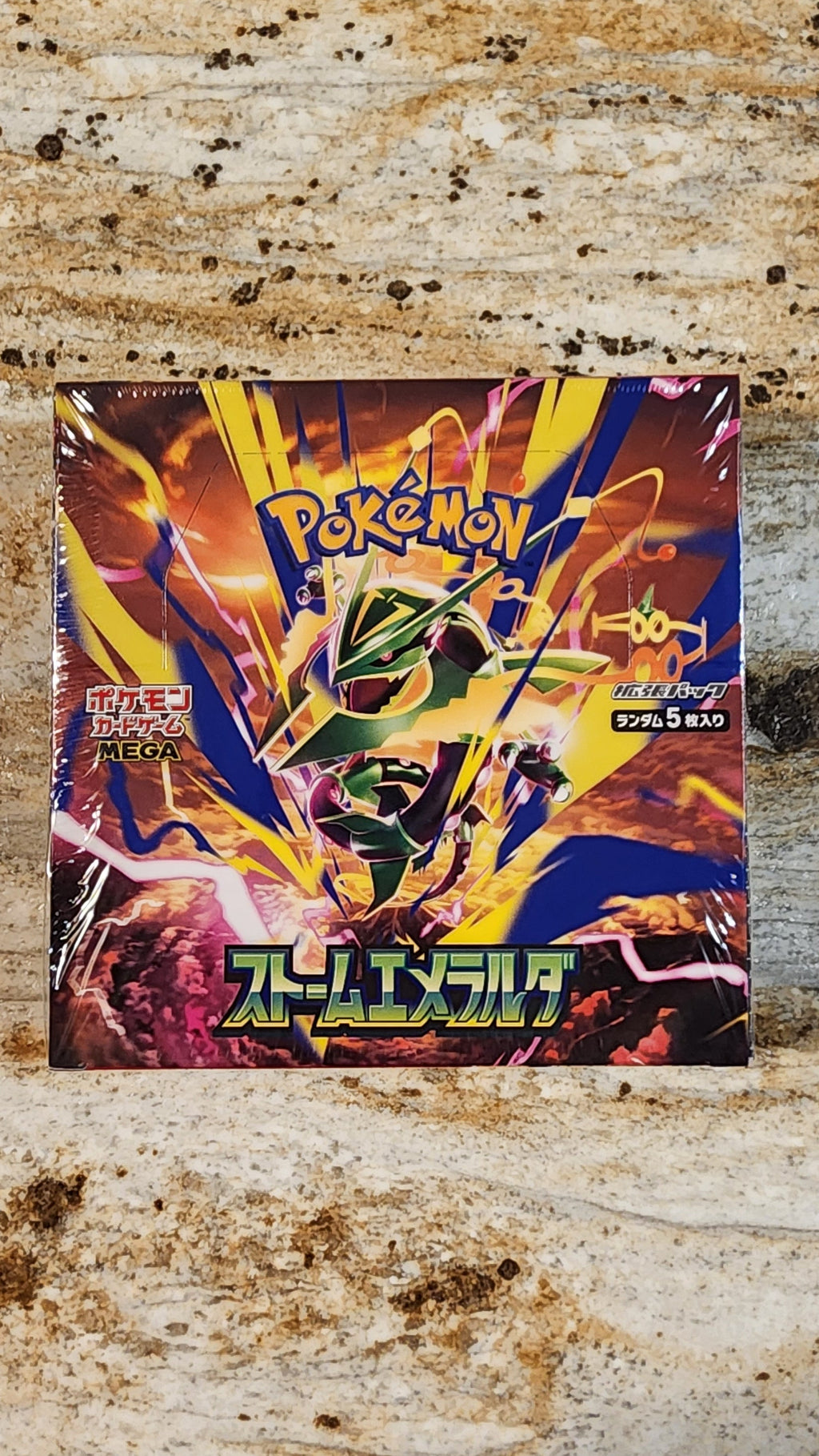 A Pokémon TCG booster box for the 2026 Storm Emeralda booster box, featuring a vibrant multicolor cover with yellow, blue, and red artwork, placed on a weathered wood surface.