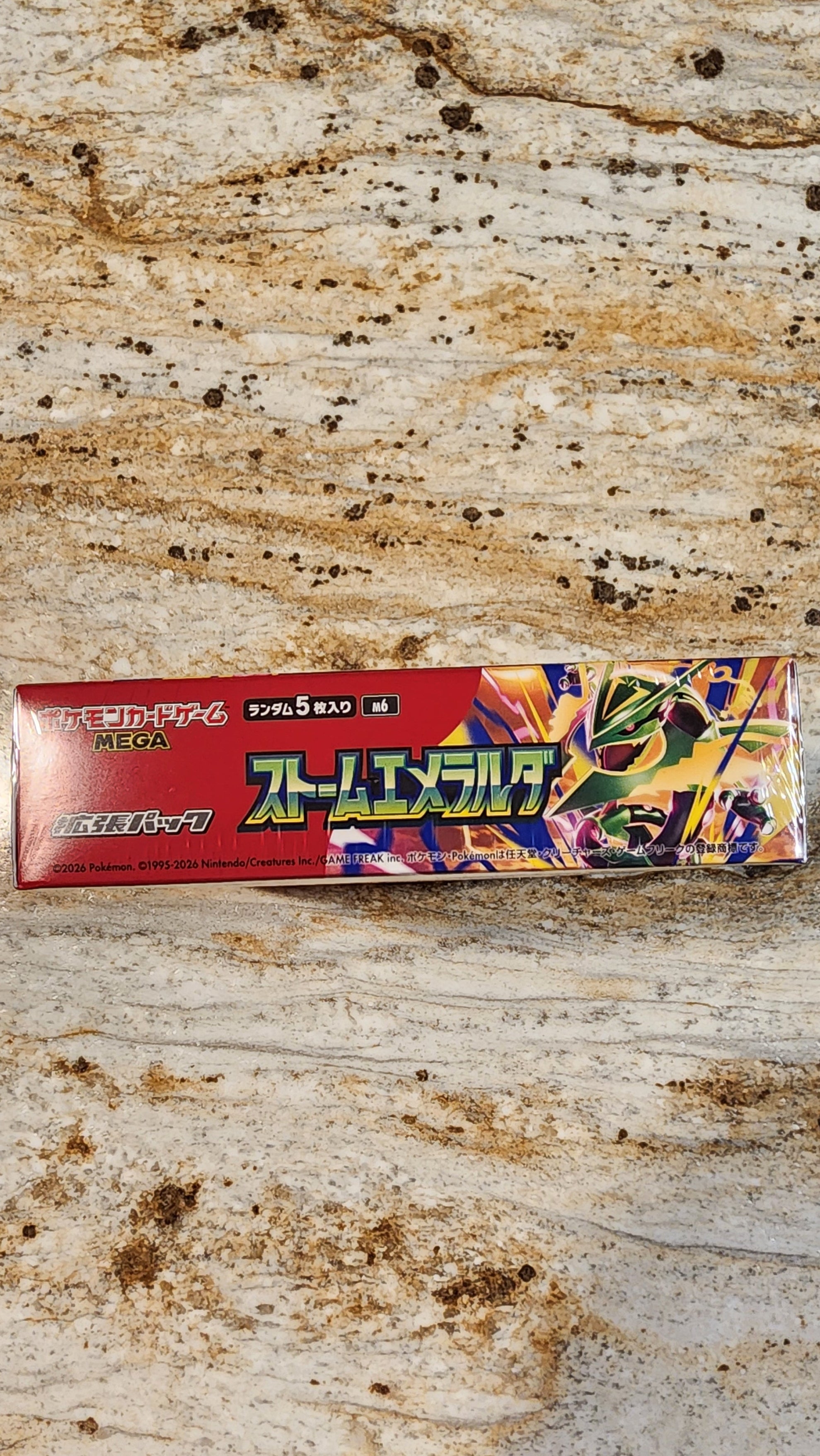 A Japanese tube of Super Glue adhesive placed on a textured, speckled stone-like surface. The packaging is primarily red with colorful graphic illustrations on the right side and prominent Japanese text.