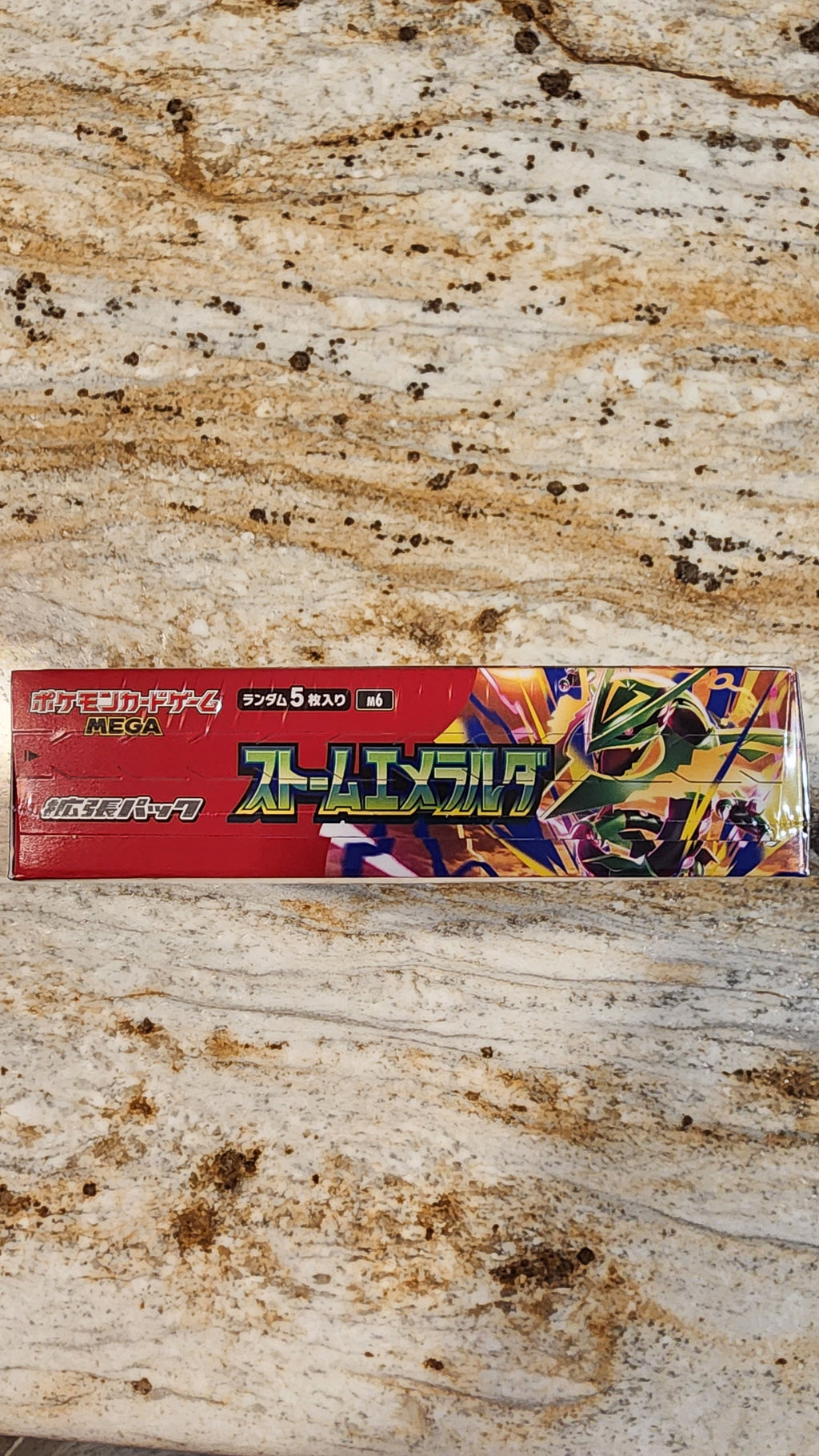 A Japanese card deck box for Pokémon resting on a heavily stained beige and brown stone tabletop.