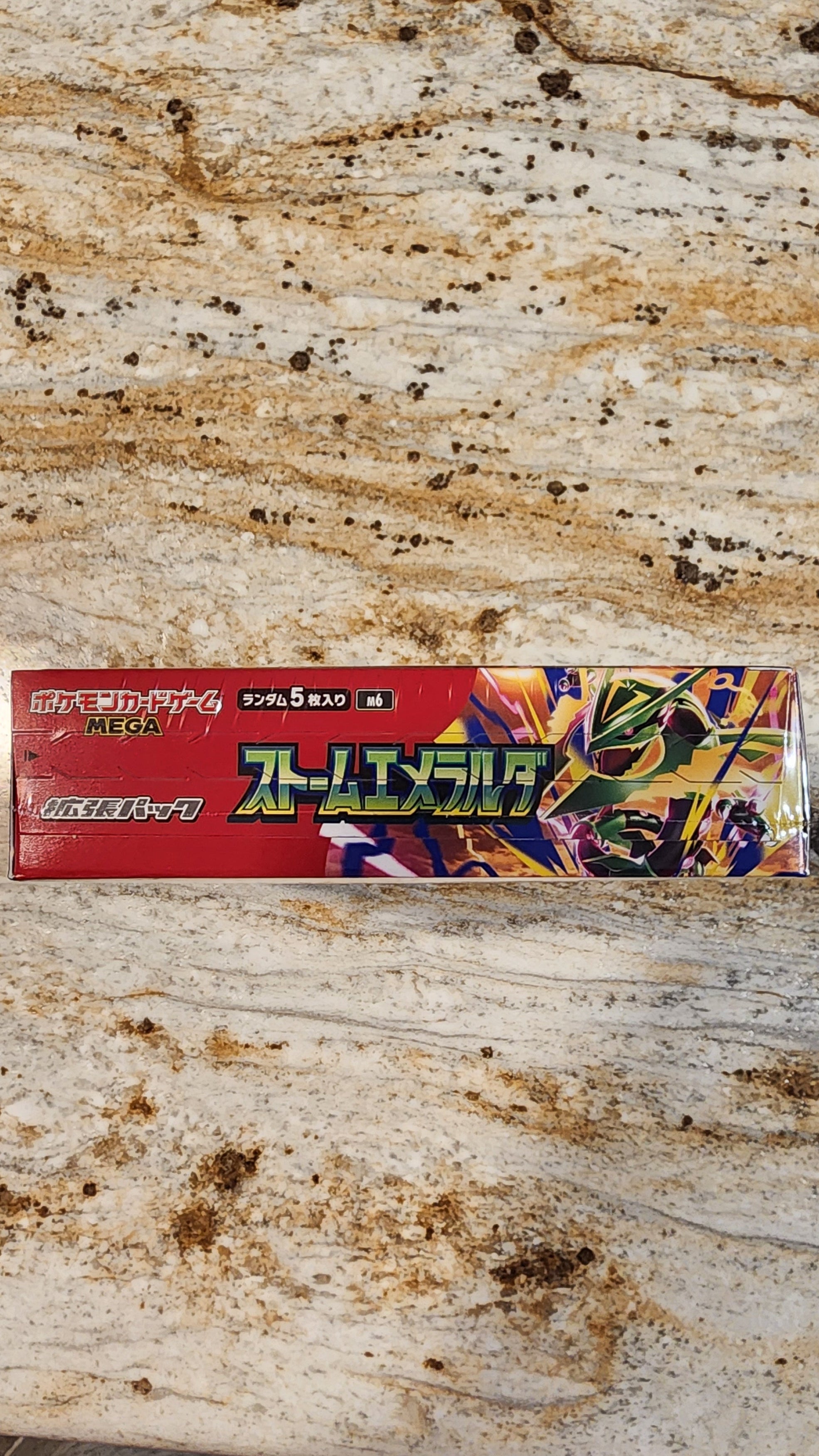A Japanese card deck box for Pokémon resting on a heavily stained beige and brown stone tabletop.