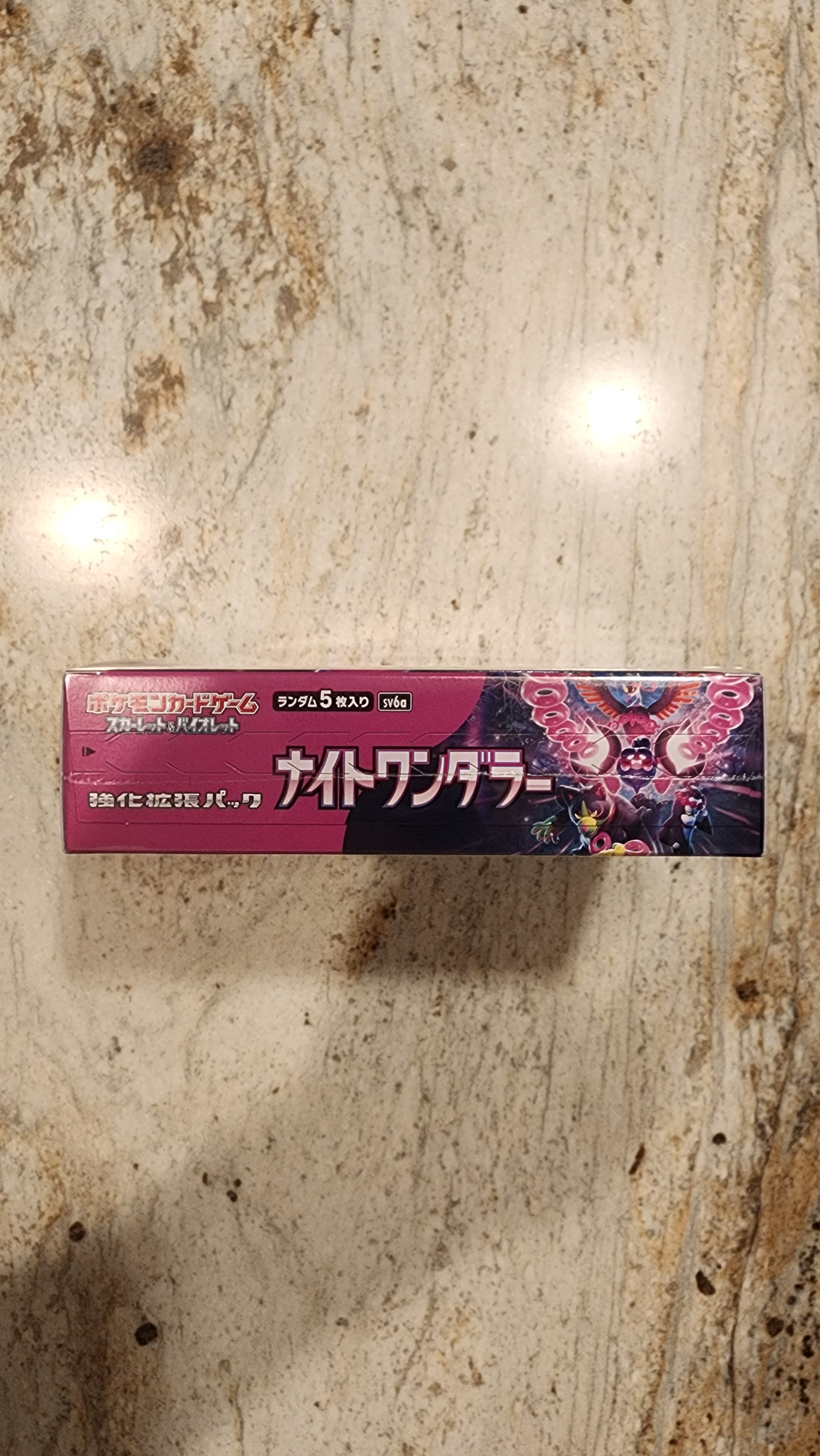 A tall rectangular product box with Japanese text and a dark pink, purple, and black design featuring a dragon illustration, standing on a beige stone surface.