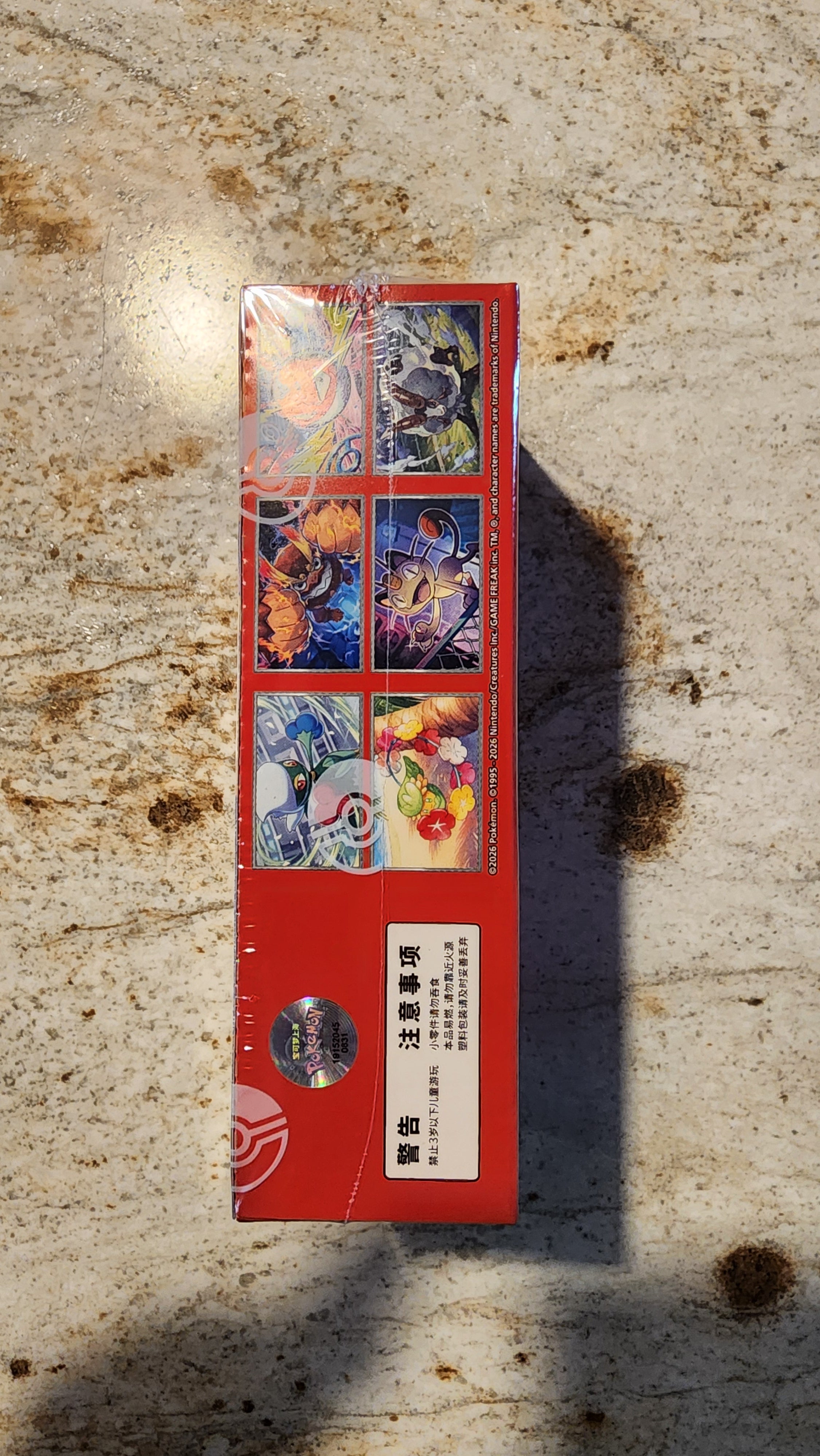 A red rectangular product box with colorful printed graphics and Chinese text, photographed standing upright on a textured beige surface with a strong shadow cast to the right.