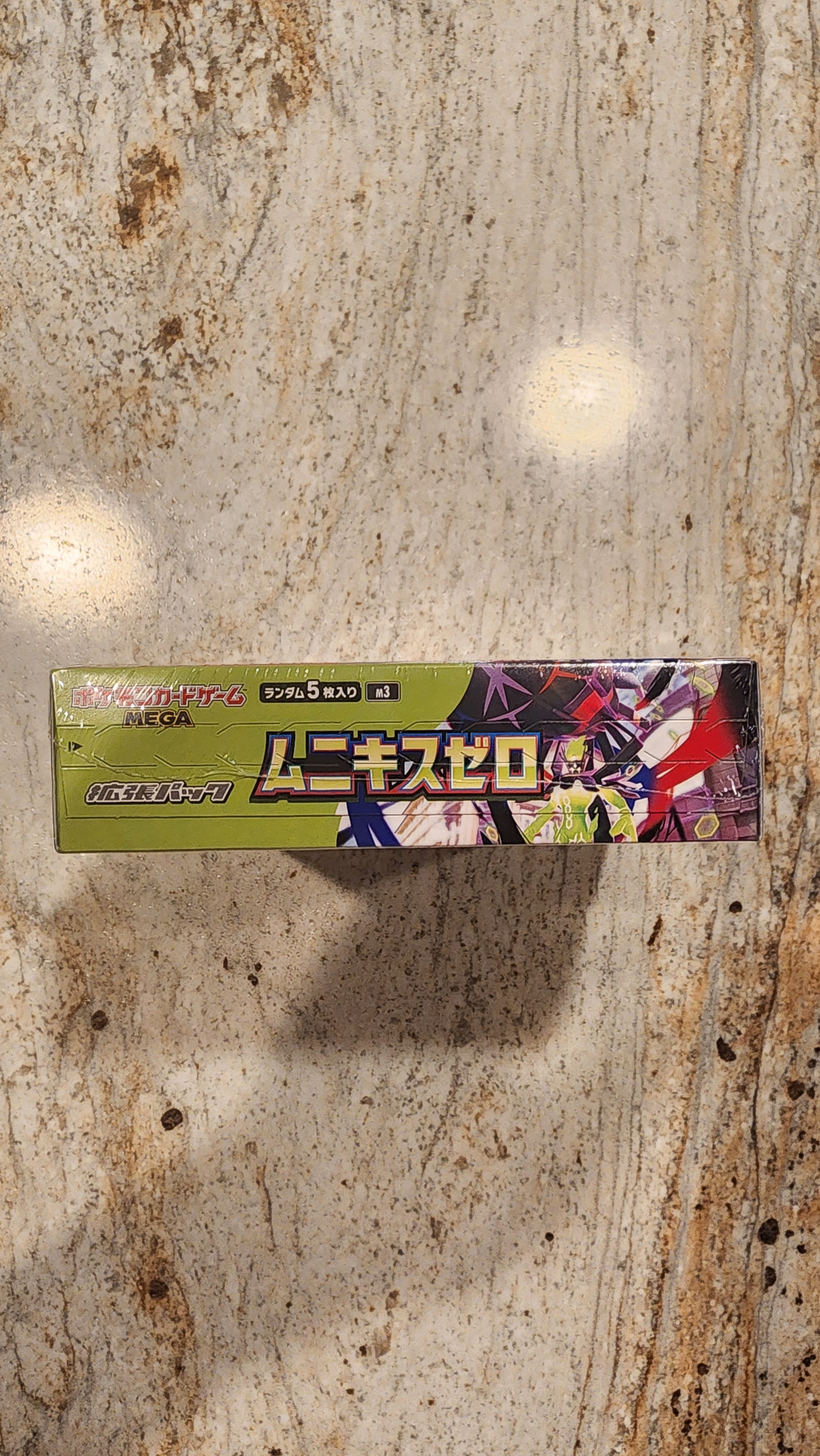 A long, narrow product box with Japanese text and colorful anime-style artwork, standing upright on a mottled beige stone surface.