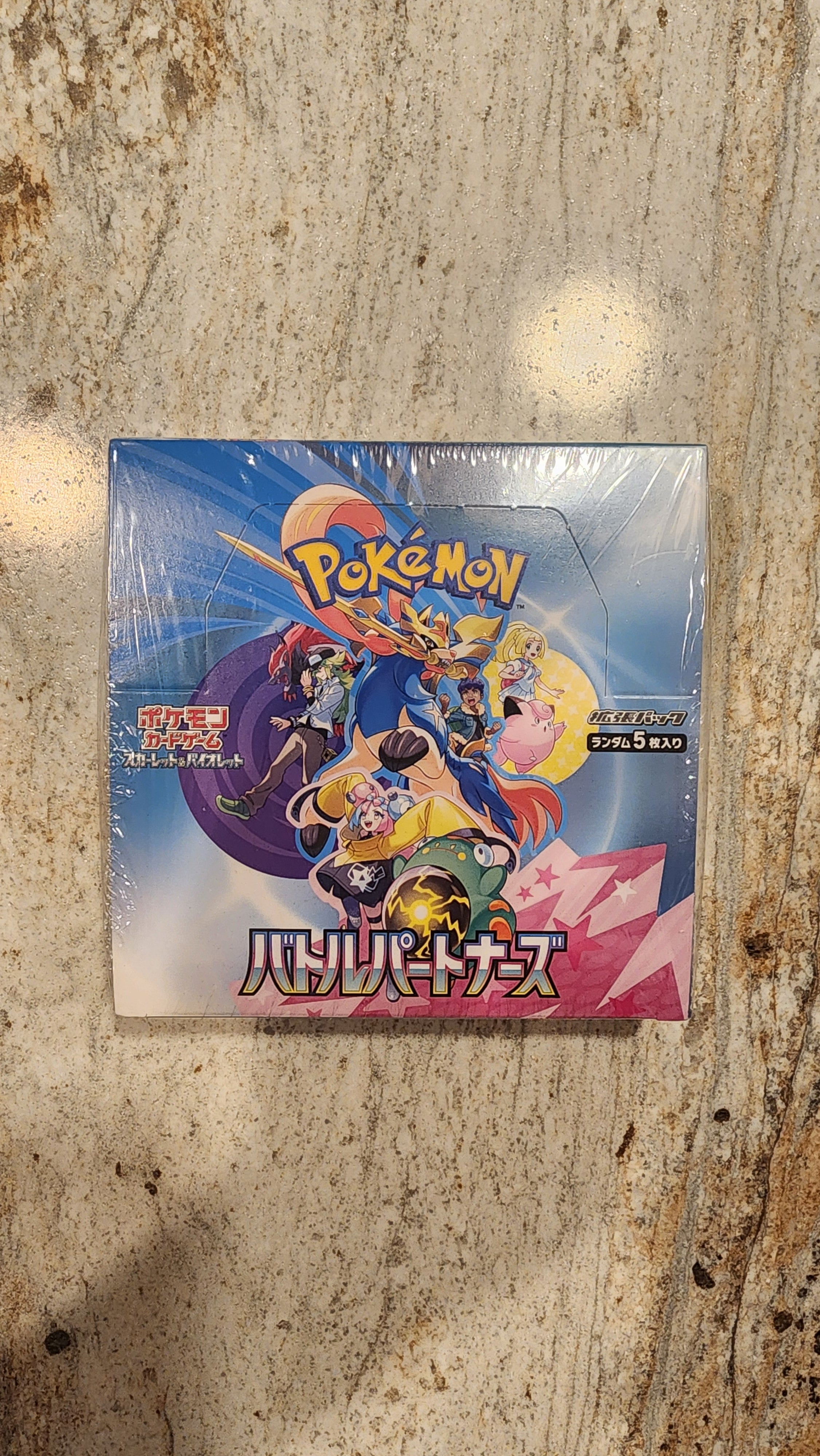 A sealed Pokémon trading card box featuring colorful anime artwork, placed on a mottled stone-like surface.