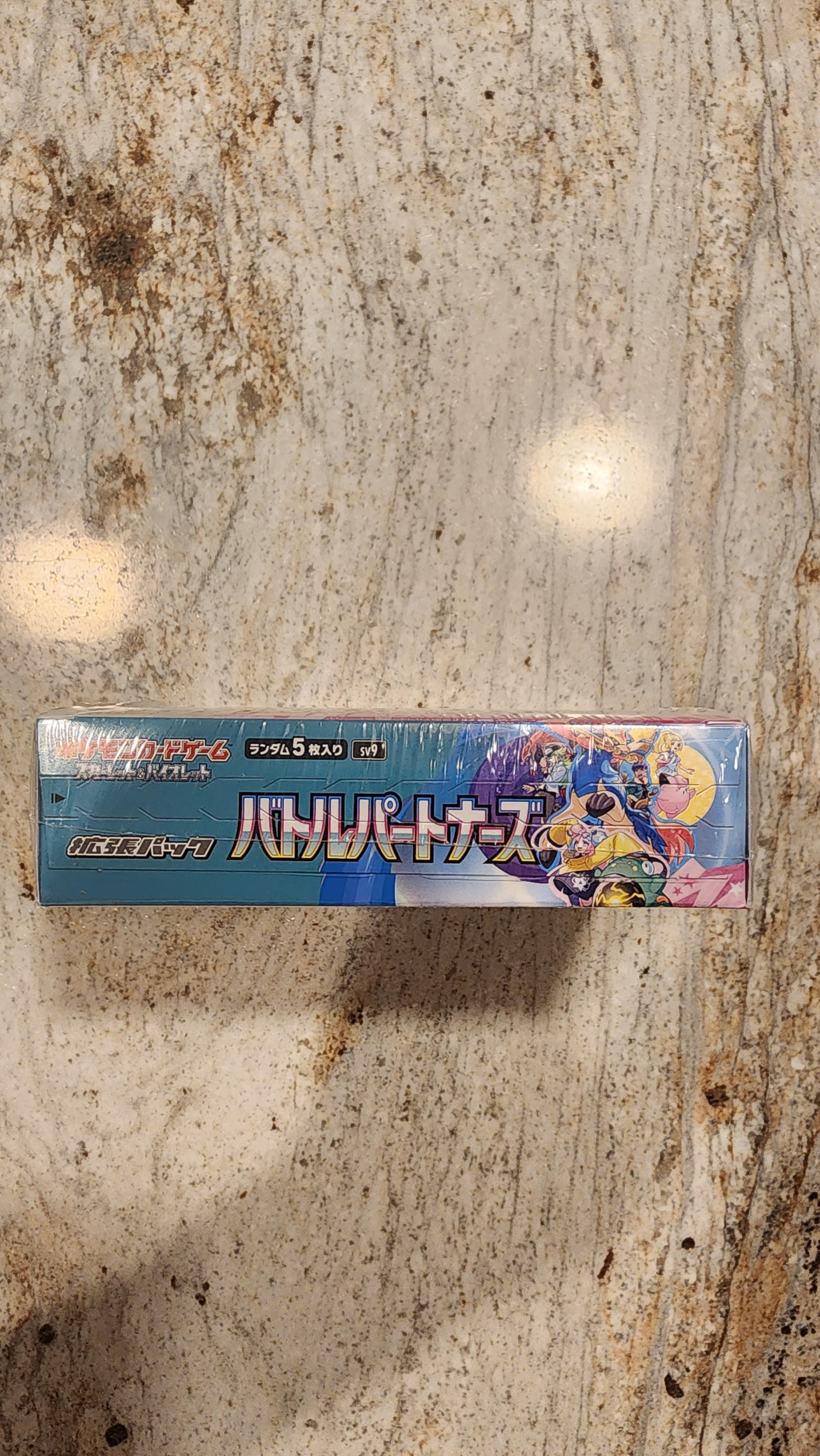 A boxed product with colorful anime-style artwork standing upright on a mottled stone surface. The package is predominantly blue with multicolored character graphics and Japanese text.