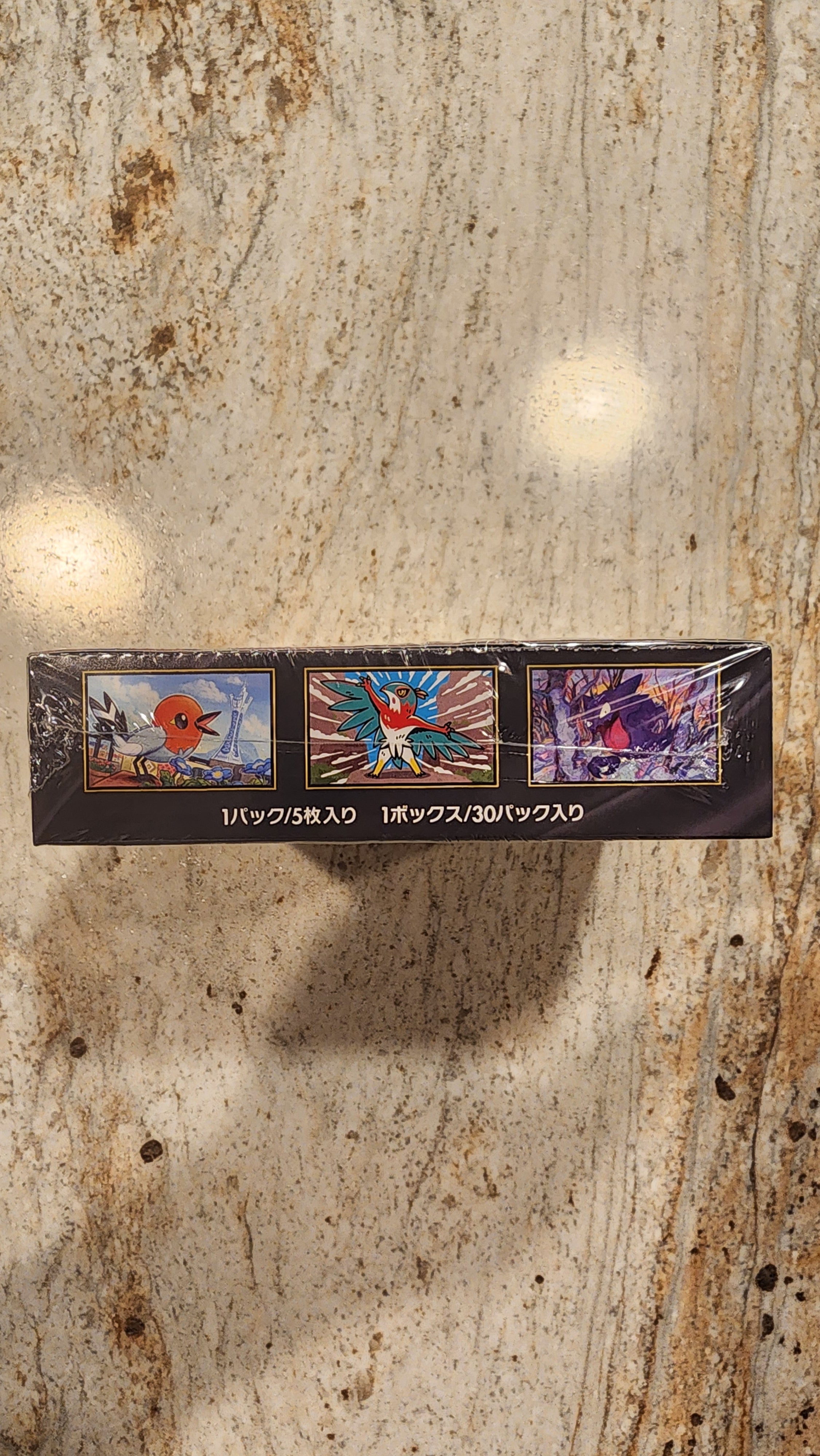 A tall, narrow black box with three colorful illustrated panels and vertical white text, photographed standing upright on a mottled beige stone surface.